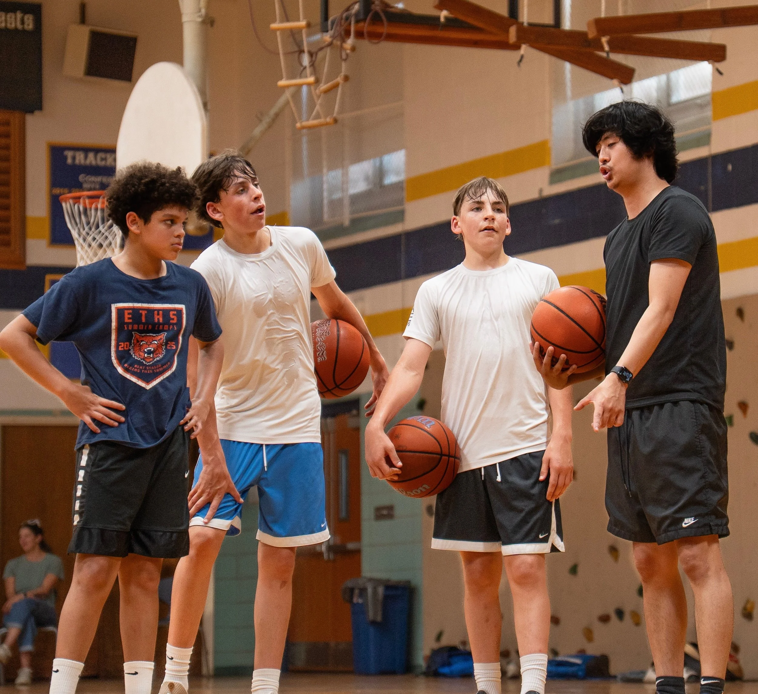 A group of four young boys standing in a gymnasium, holding basketballs, and engaged in conversation during a basketball practice or game. They are wearing athletic clothing, and a basketball hoop and climbing wall are visible in the background.