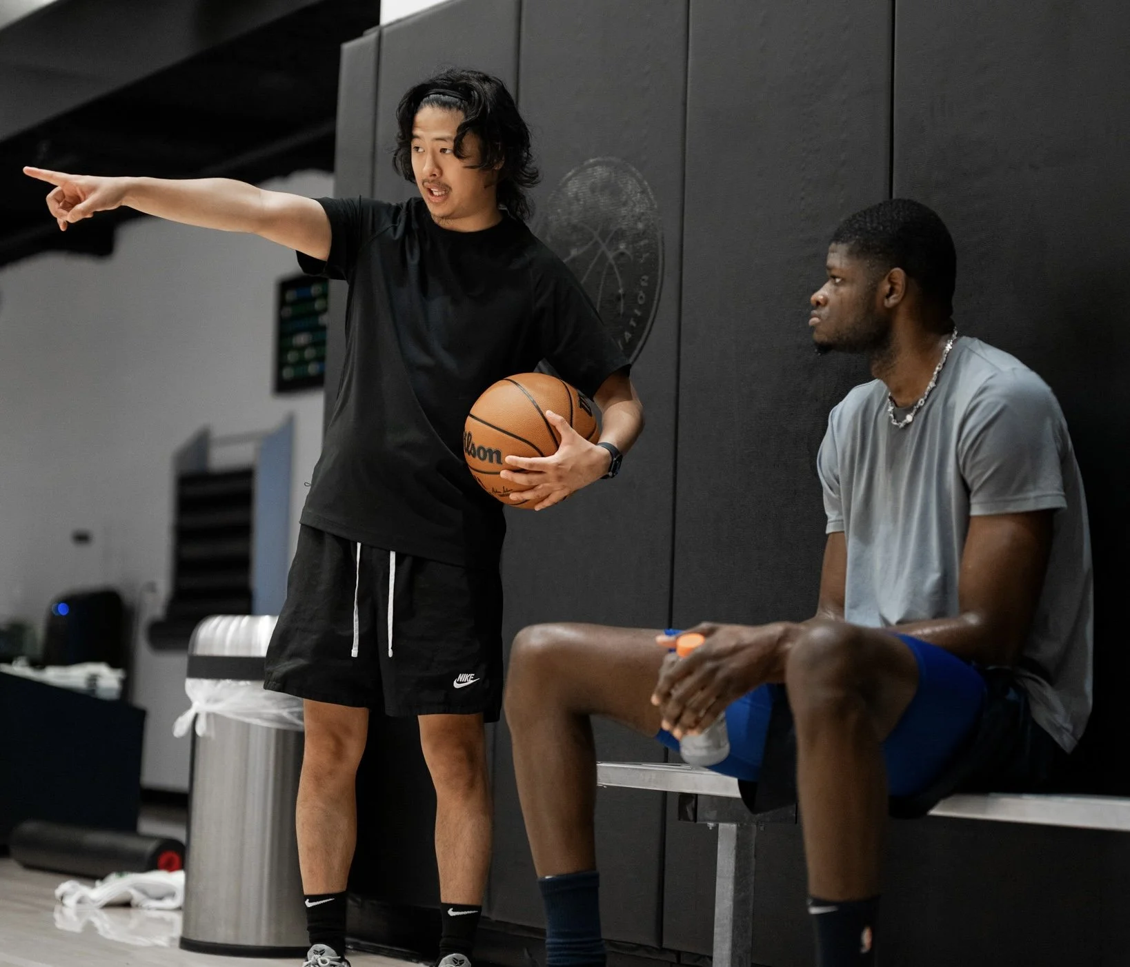 A man with long dark hair holding a basketball points while talking to a seated man in a gym locker room.