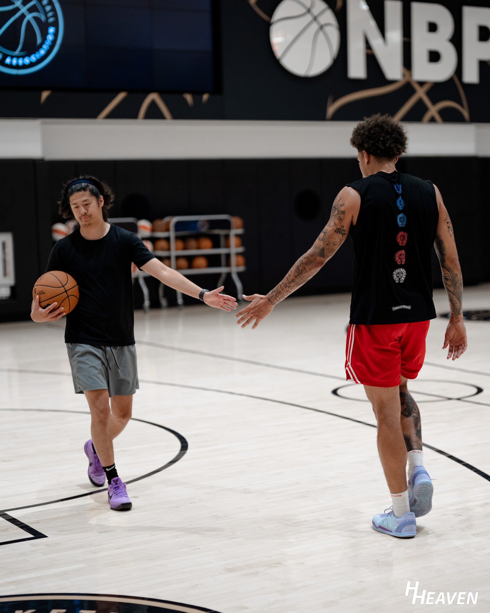 Two young men practicing basketball in an indoor court, one of them holding a basketball, with basketballs and basketball hoops in the background.