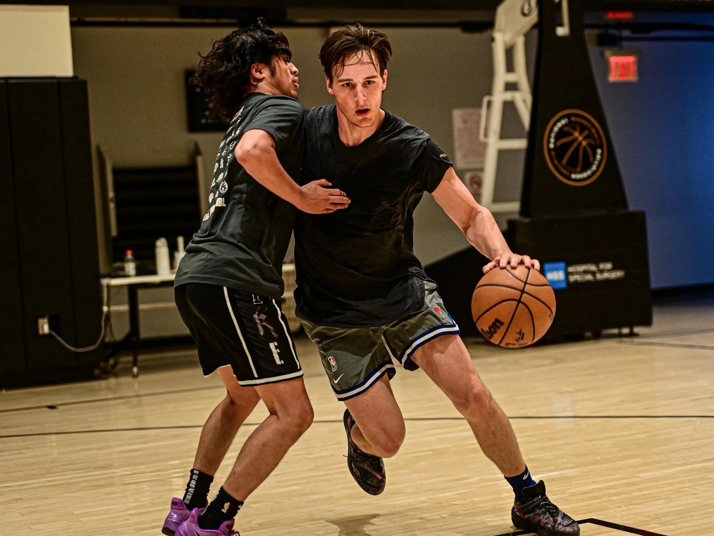 Two young men playing basketball on an indoor court. One is dribbling the ball while the other is attempting to block him. Both are focused and in motion.