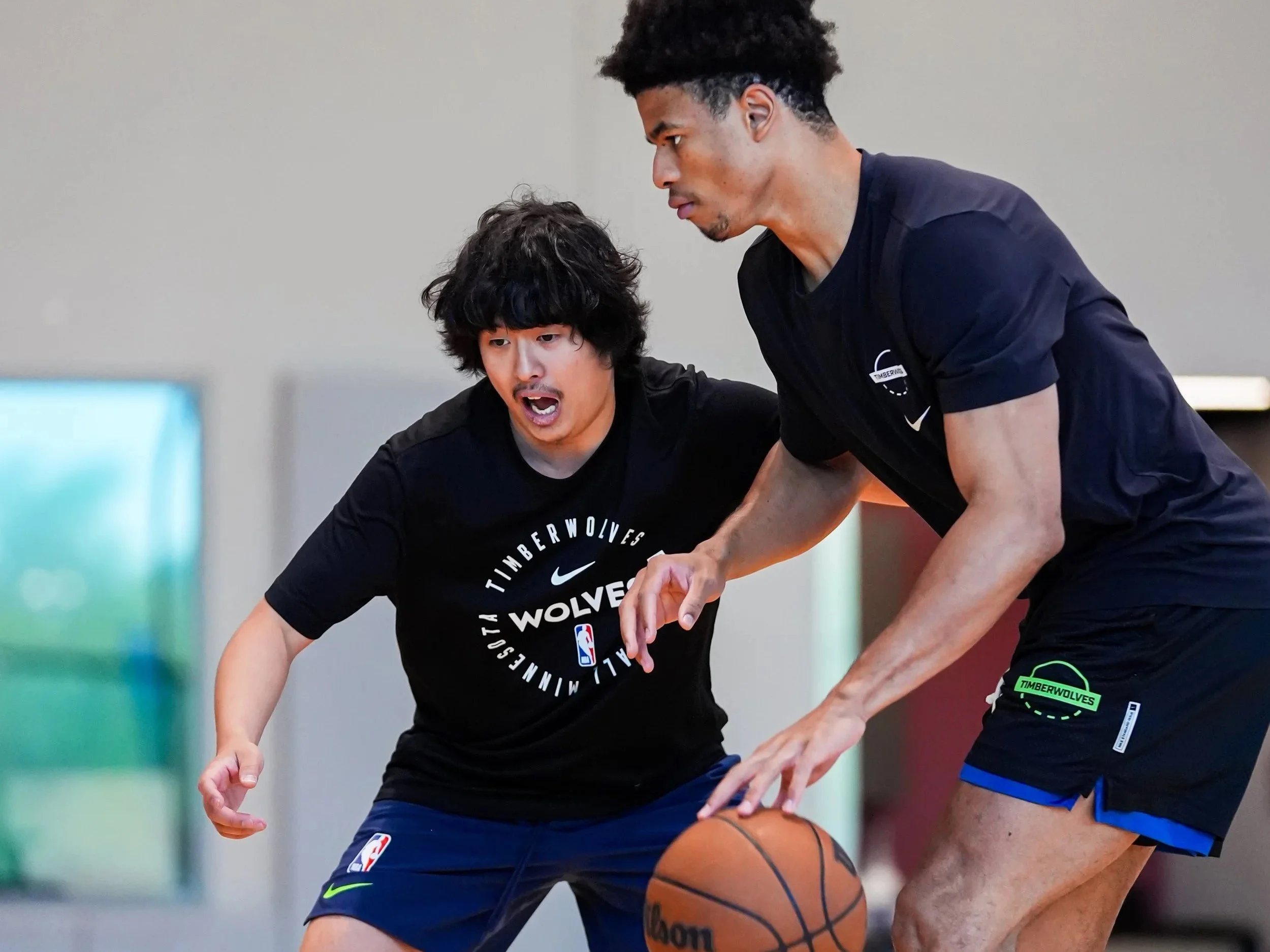 Two basketball players practice dribbling during a game or training session, with a plain background.