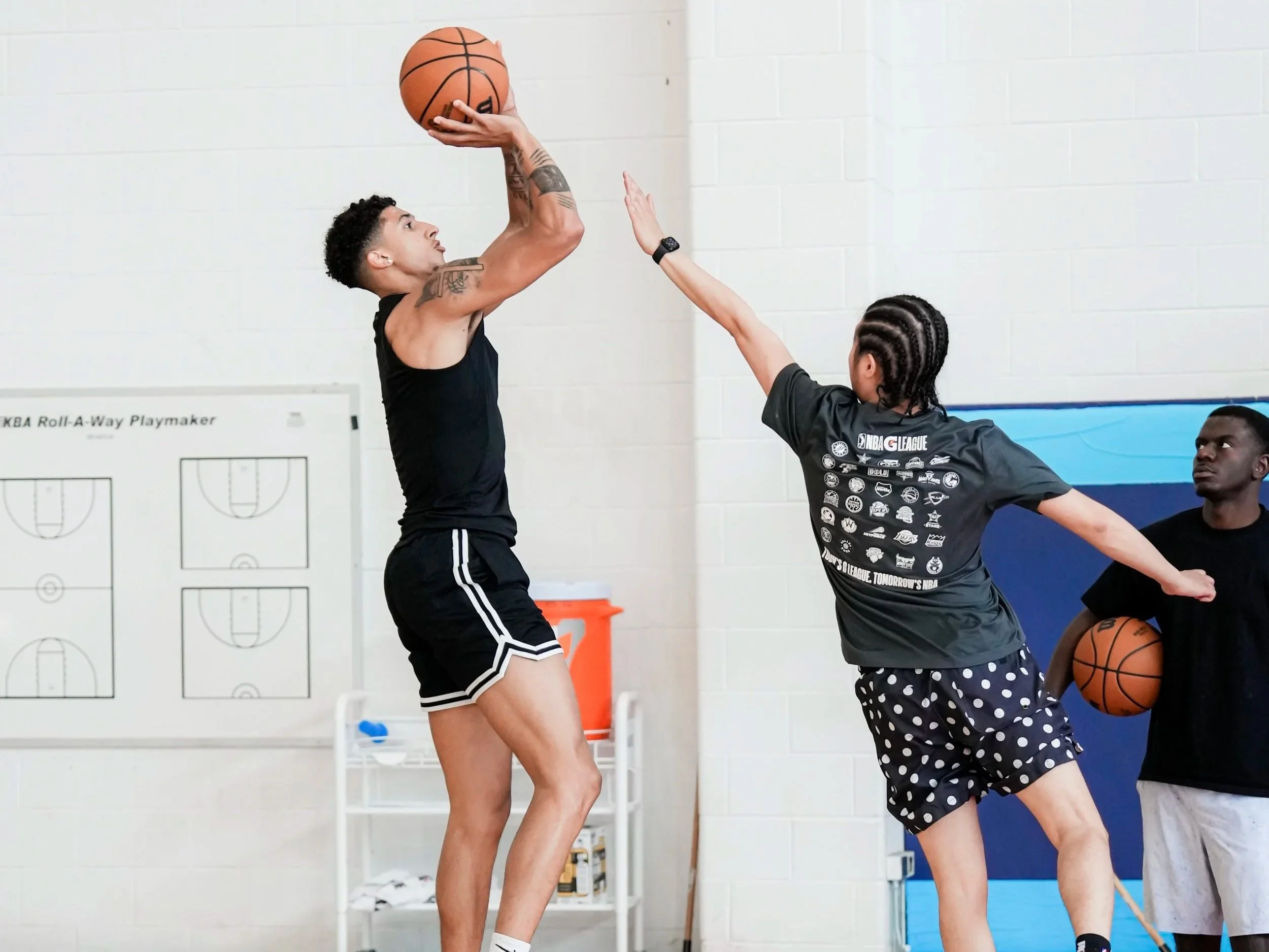 A person holding a basketball above their head while another person attempts to block or defend during a basketball game or practice. The players are indoors, with white brick walls in the background and a basketball court visible.