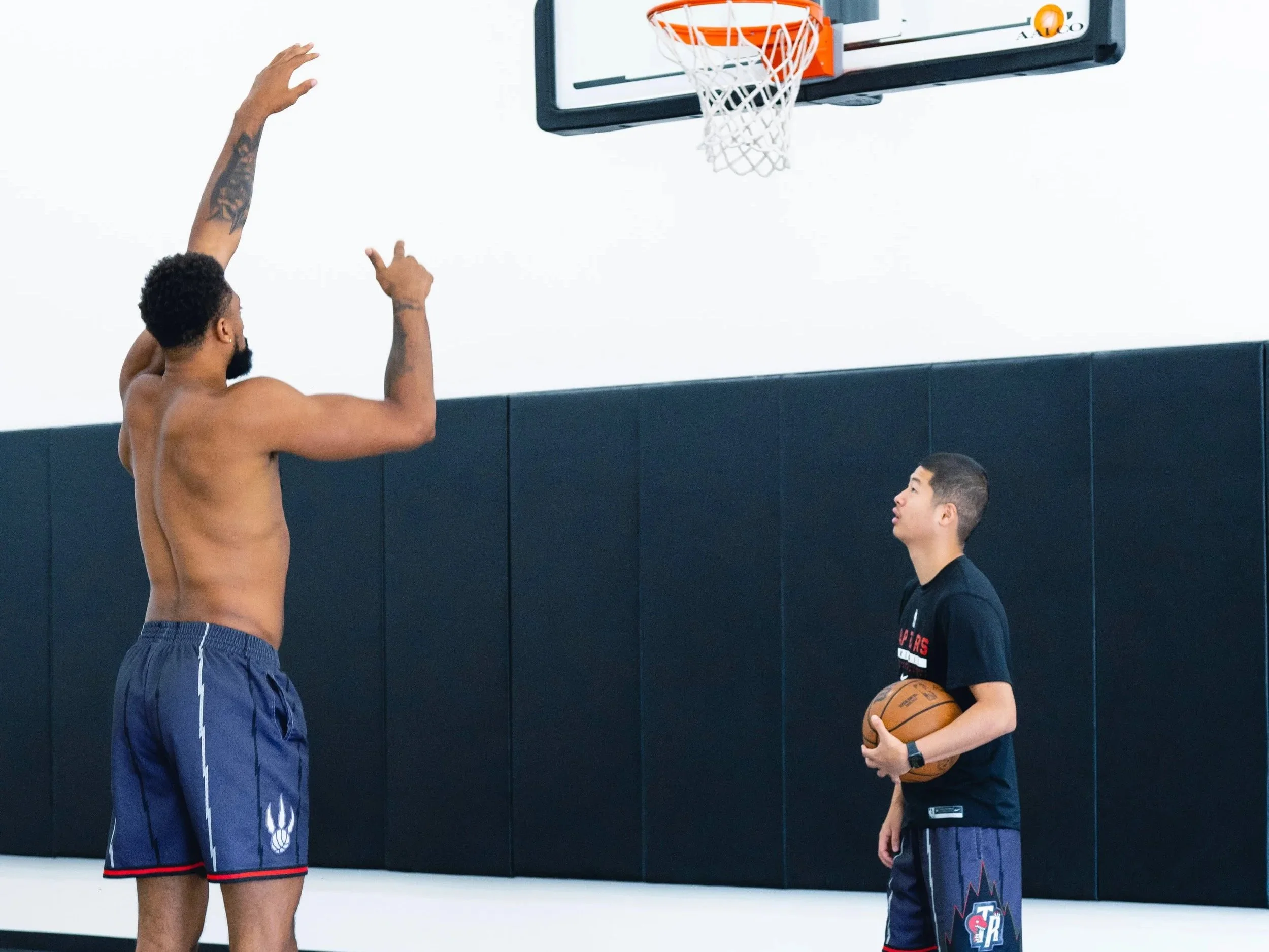 A shirtless man preparing to shoot a basketball while a young boy holding a basketball and wearing a black T-shirt and shorts watches him in a gym with a basketball hoop in the background.
