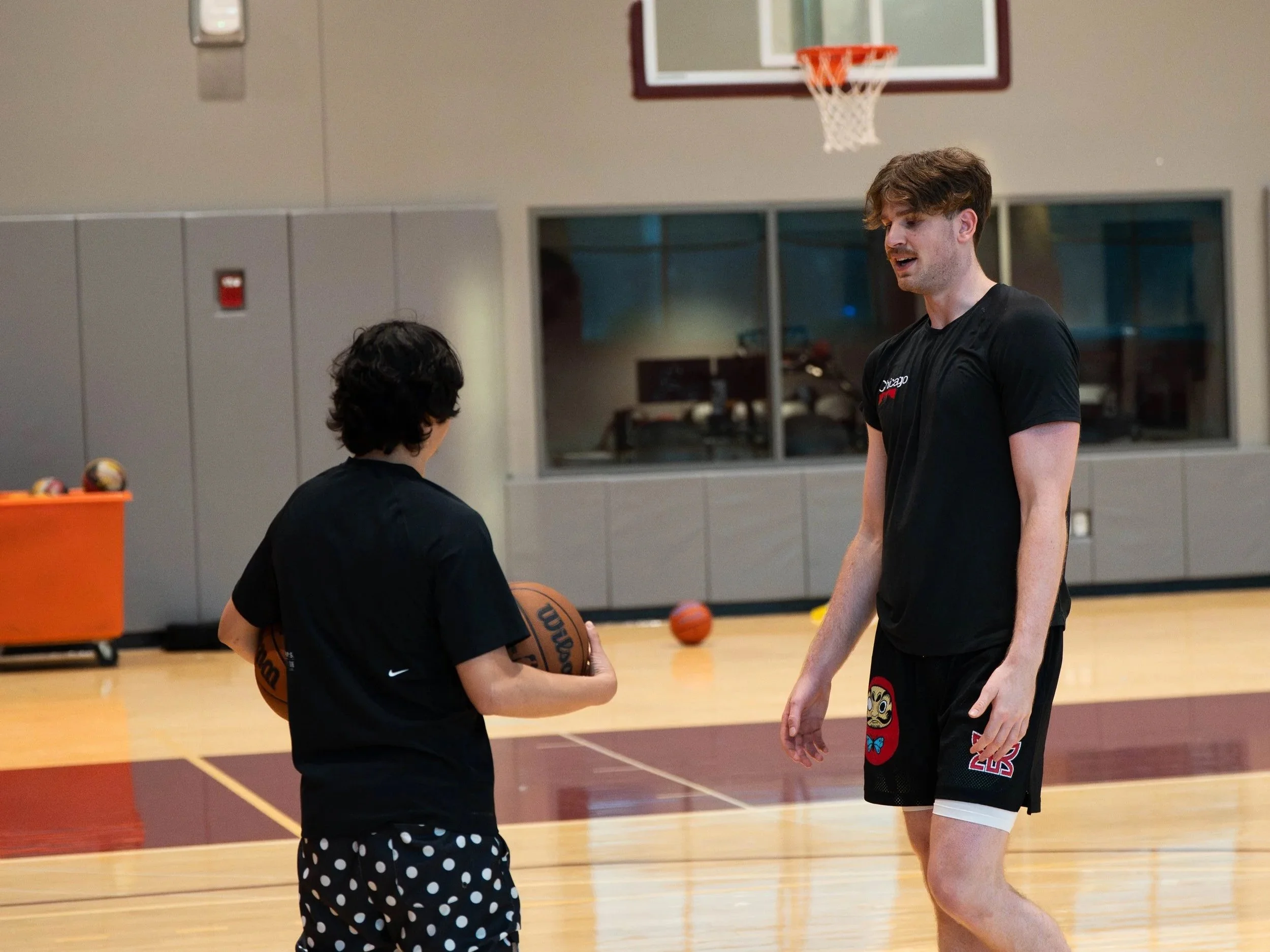 A young boy with dark hair holding a basketball and talking to an adult male basketball player in a gymnasium.