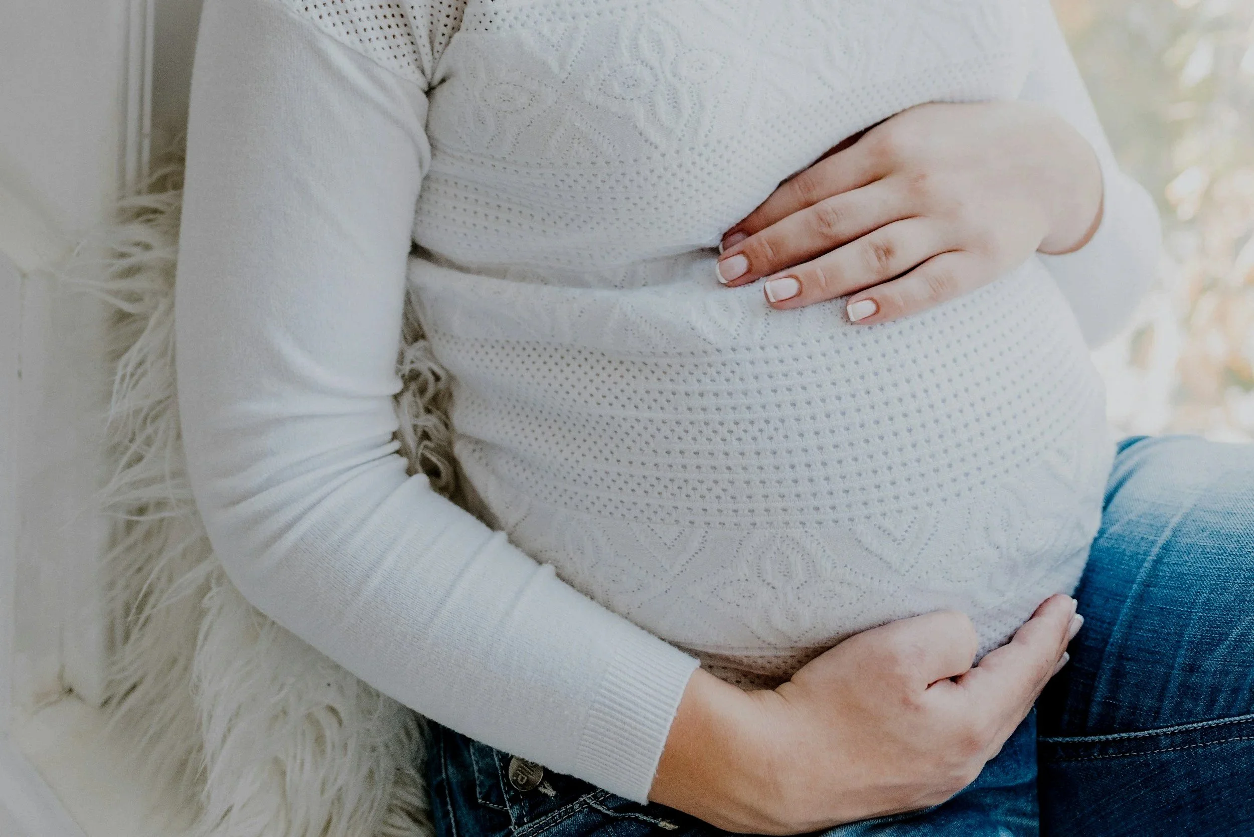 A pregnant woman wearing a white long-sleeve shirt with her hand resting on her belly, sitting on a fluffy white surface. She is in natural light, with a soft background.