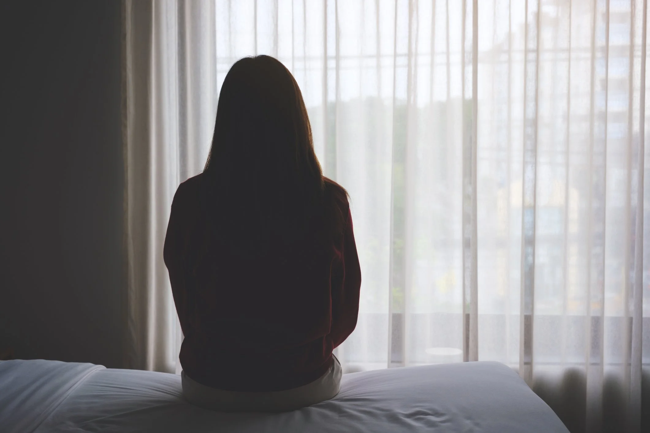 A woman sitting on a bed facing a window with sheer curtains, back viewed
