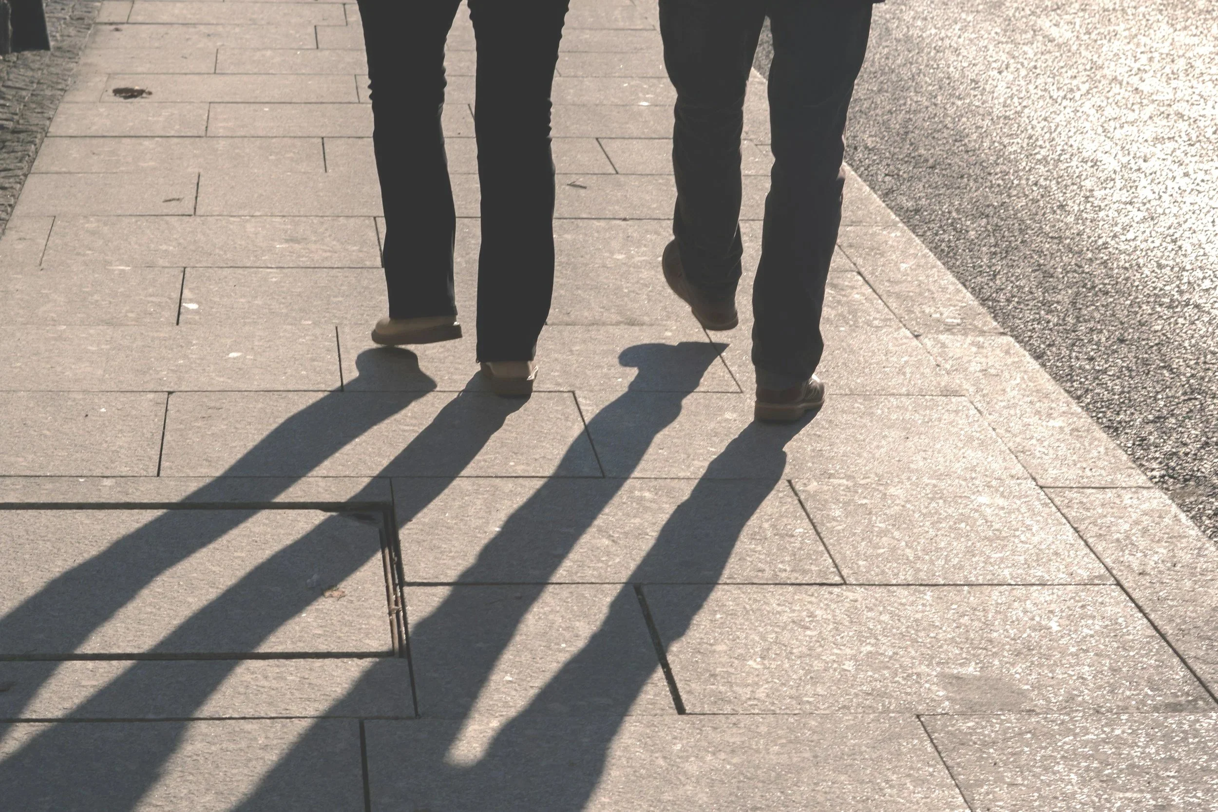 Two people walking on a sidewalk with their shadows cast on the ground. The image shows only their legs and feet, and the sun is shining brightly, creating distinct shadows.