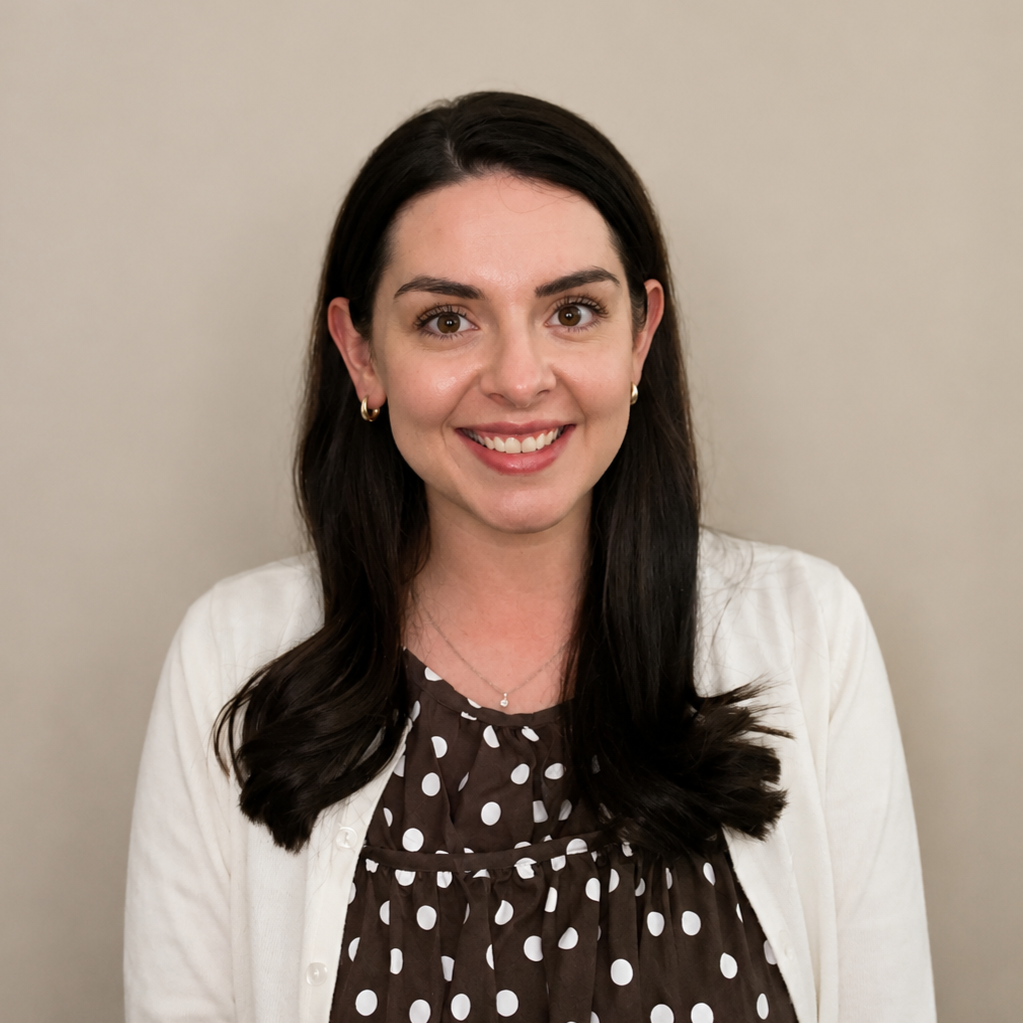 A woman with long dark hair and hoop earrings smiling at the camera, wearing a white cardigan and a brown polka dot blouse, against a plain beige background.