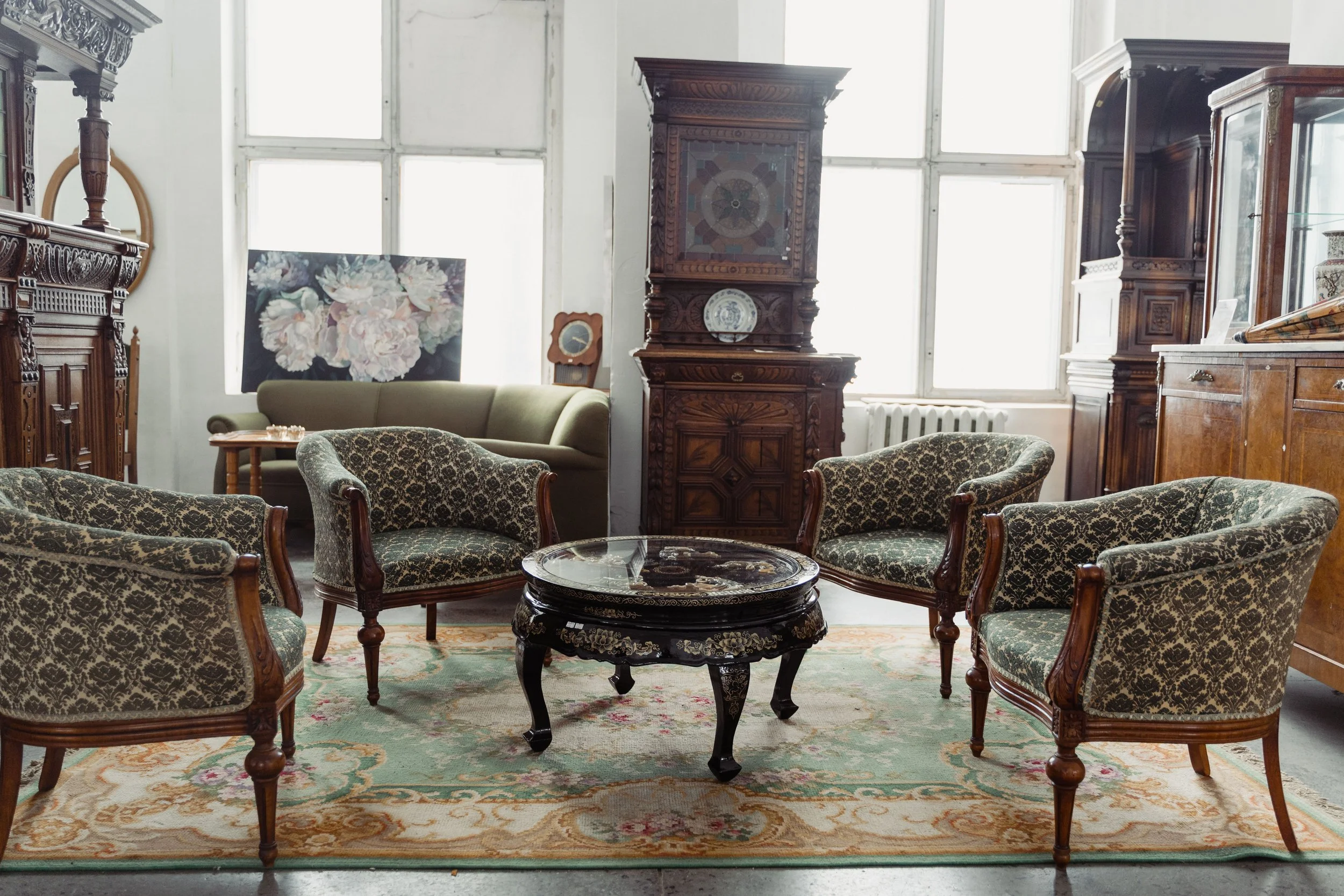 A room filled with vintage furniture including a sofa, armchairs, and several wooden cabinets and shelves. There is a painting of large flowers on the wall, several windows providing natural light, and a carpet covering the floor.