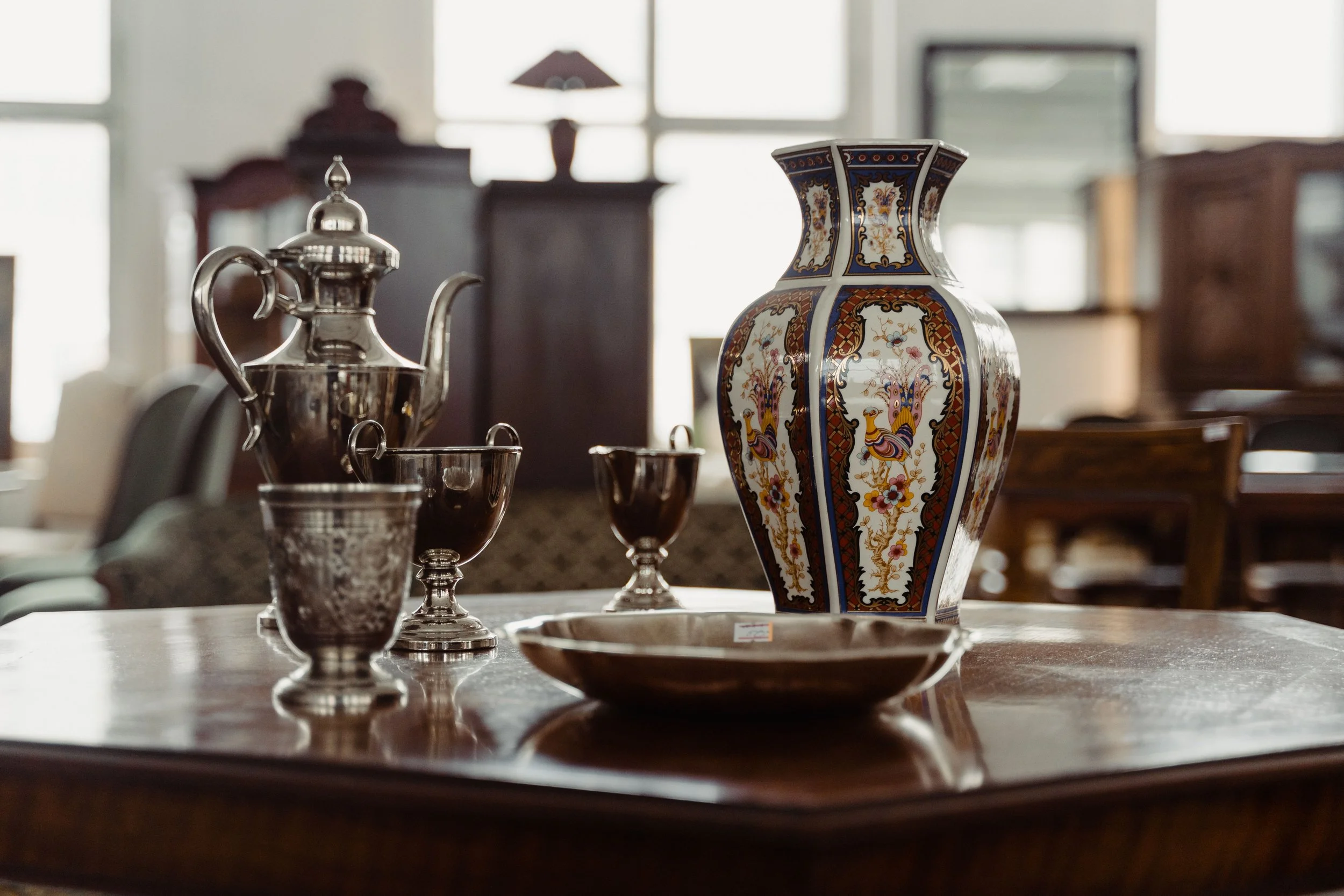 An assortment of decorative items on a wooden table, including a colorful porcelain vase, a silver teapot, and silver cups, with blurred furniture and windows in the background.