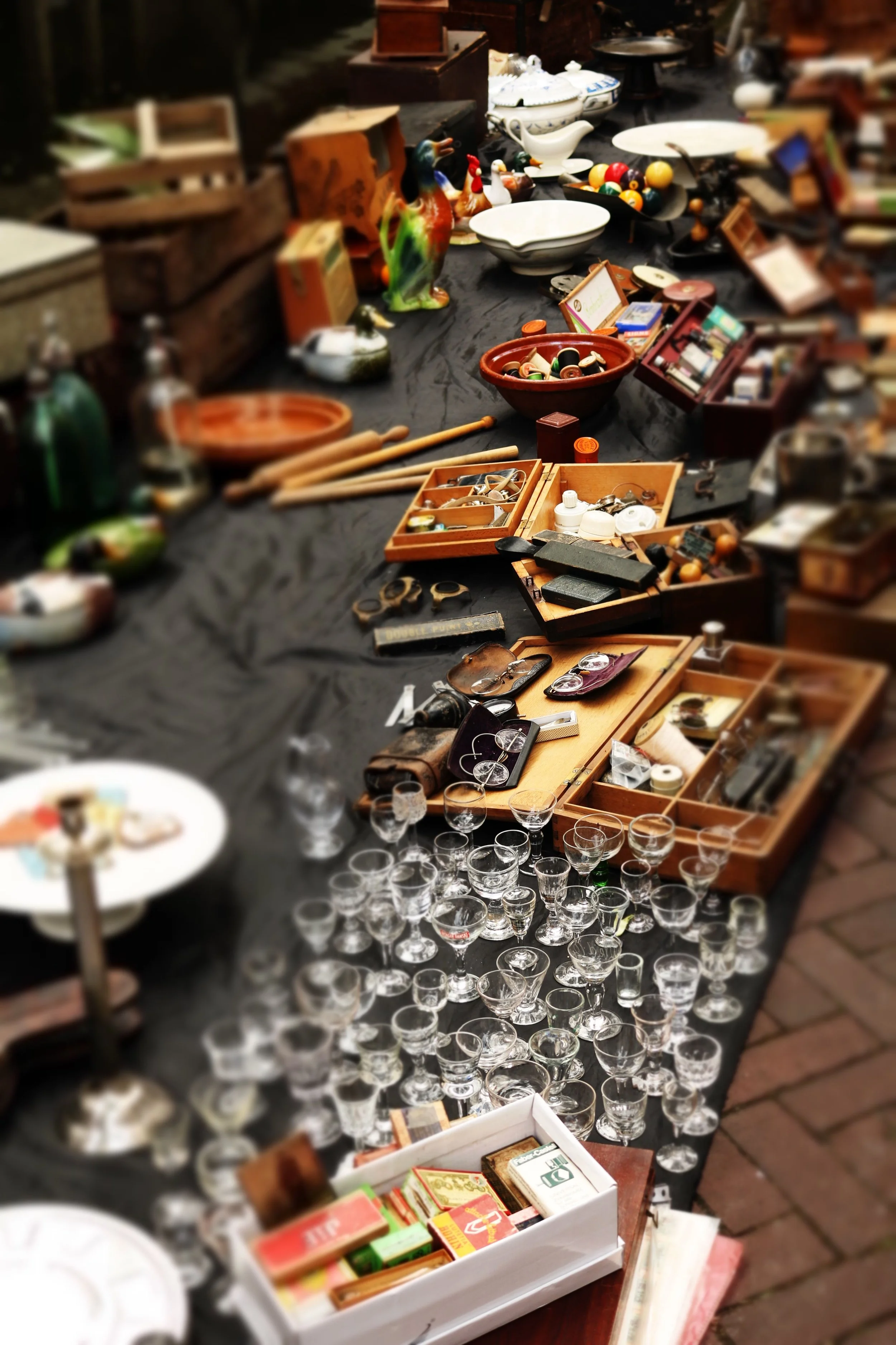 A cluttered table at a flea market with various ceramic, glassware, and boxed items, including a colorful ceramic duck, bowls, kitchen utensils, and small collectibles.