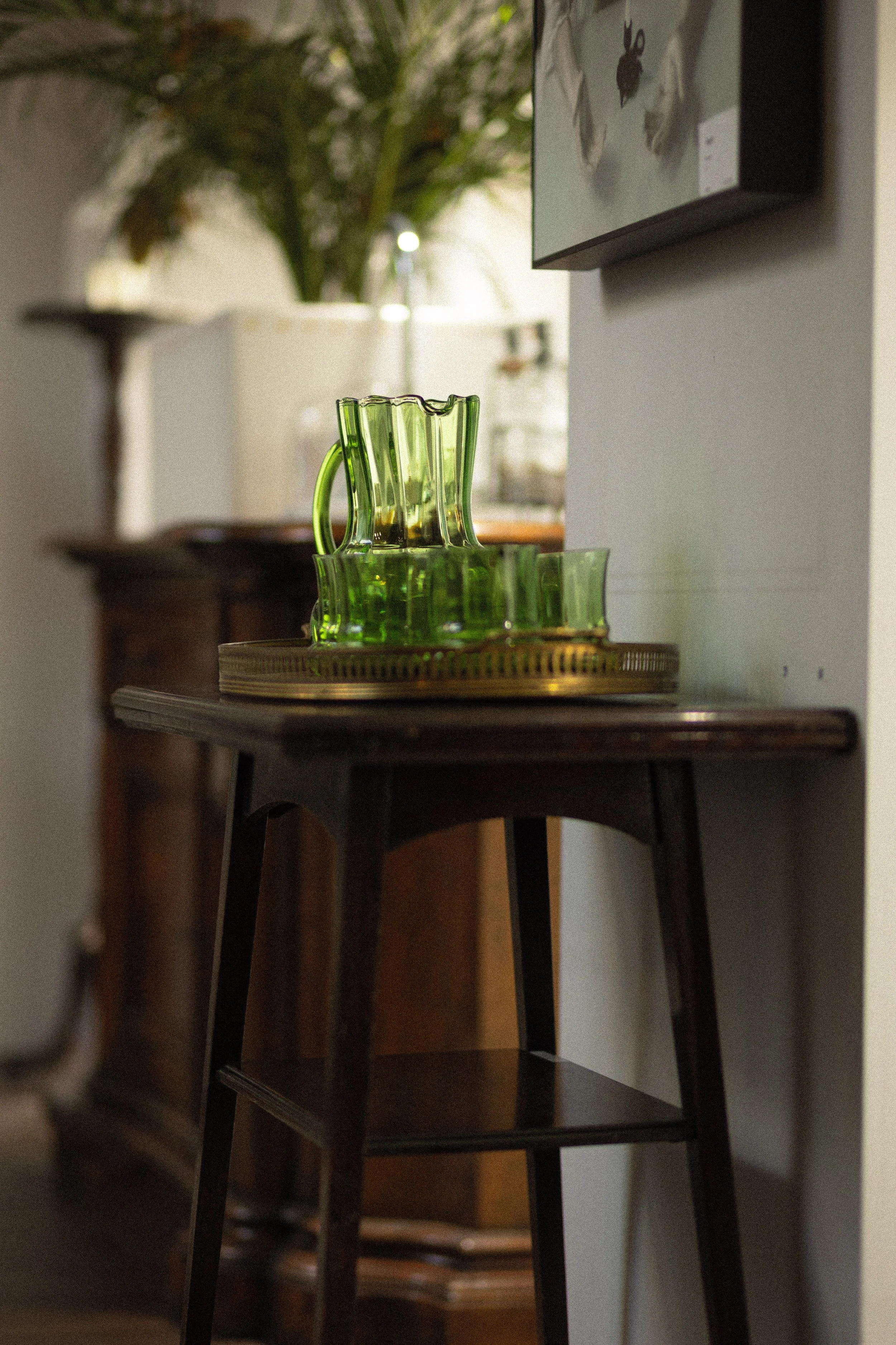 A close-up of a wooden table with a brass tray holding a nested set of green glass pitchers, with a blurred background of a houseplant and a wall-mounted picture.