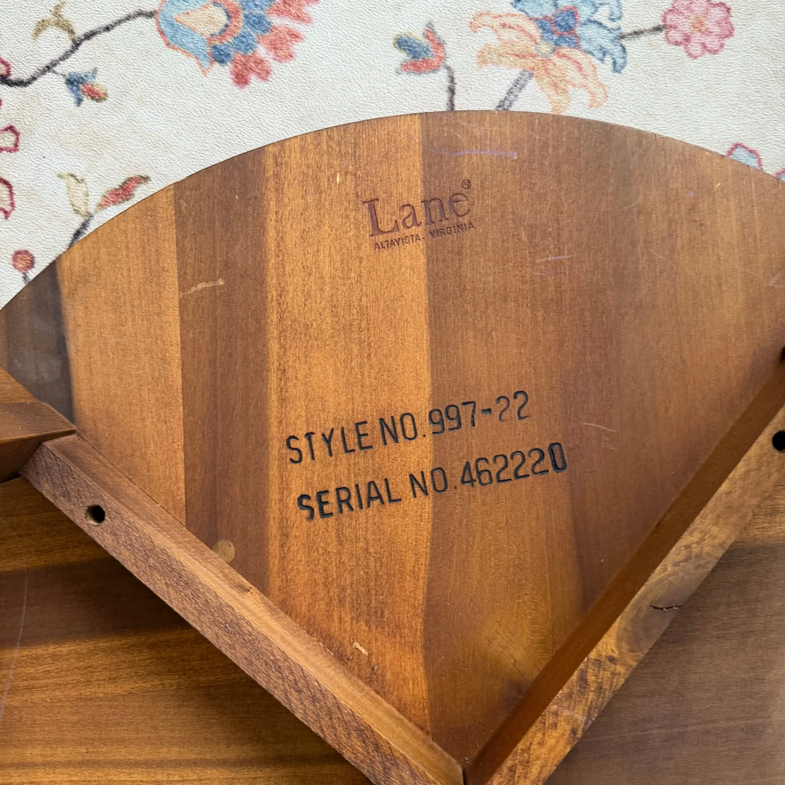 Close-up of a circular wooden object with the words 'Lane' and 'Altavista, Virginia' engraved, placed on a wood surface, with part of a floral-patterned cloth in the background.