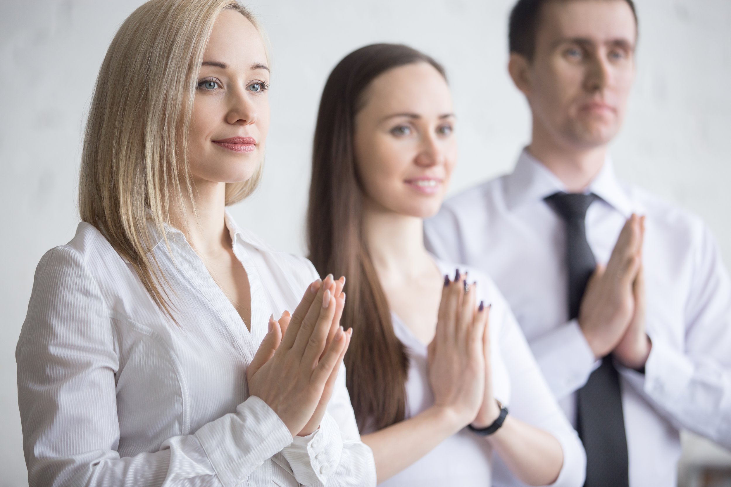 Three young corporate professionals in business attire practicing a mindful meditation prayer gesture, showcasing workplace wellness and office yoga programs for hybrid teams in the Norwest and Hills District regions.