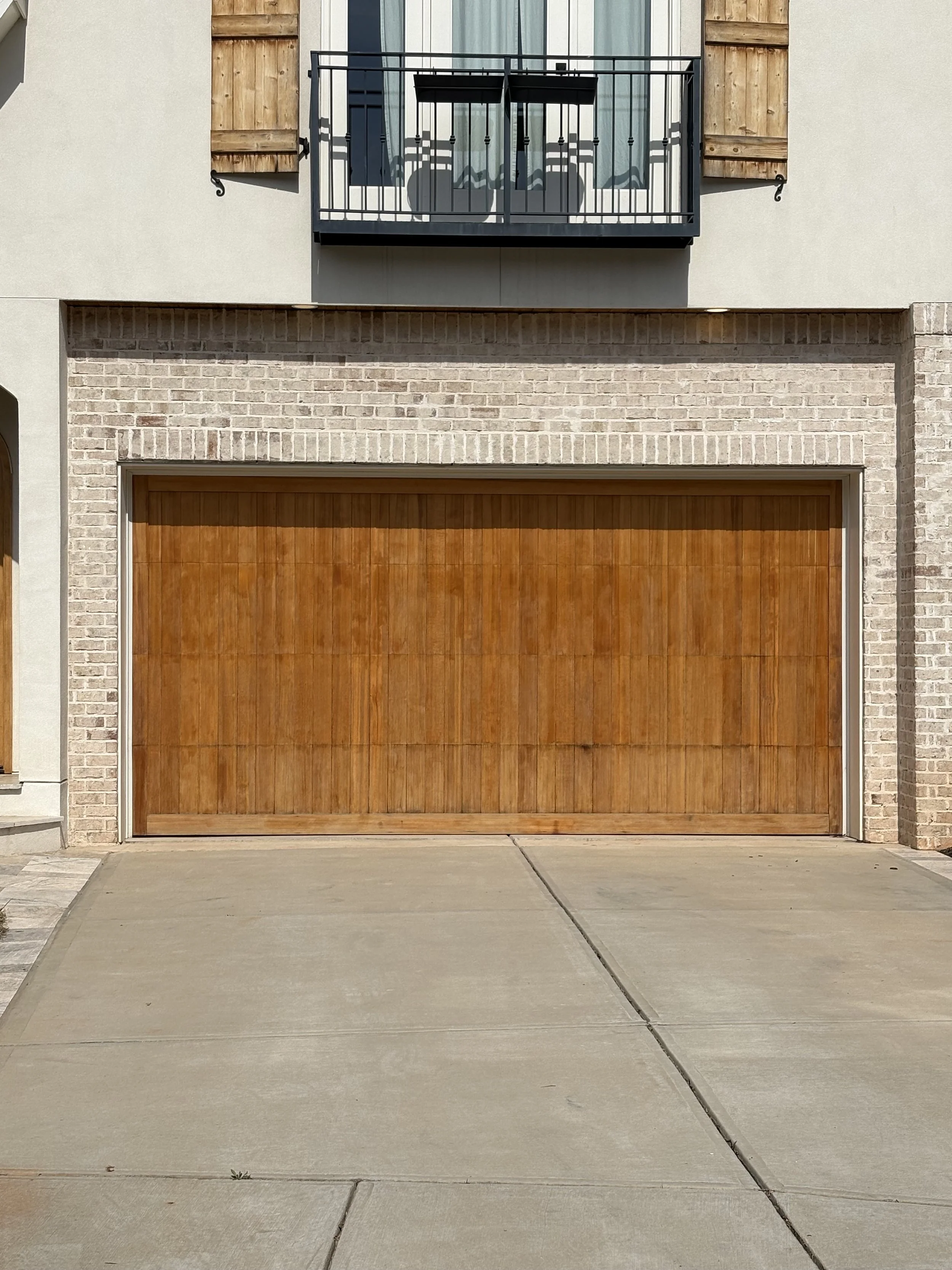 Front view of a house with a concrete driveway, a wooden garage door, brick and stucco exterior walls, and a small balcony with metal railing above the garage.
