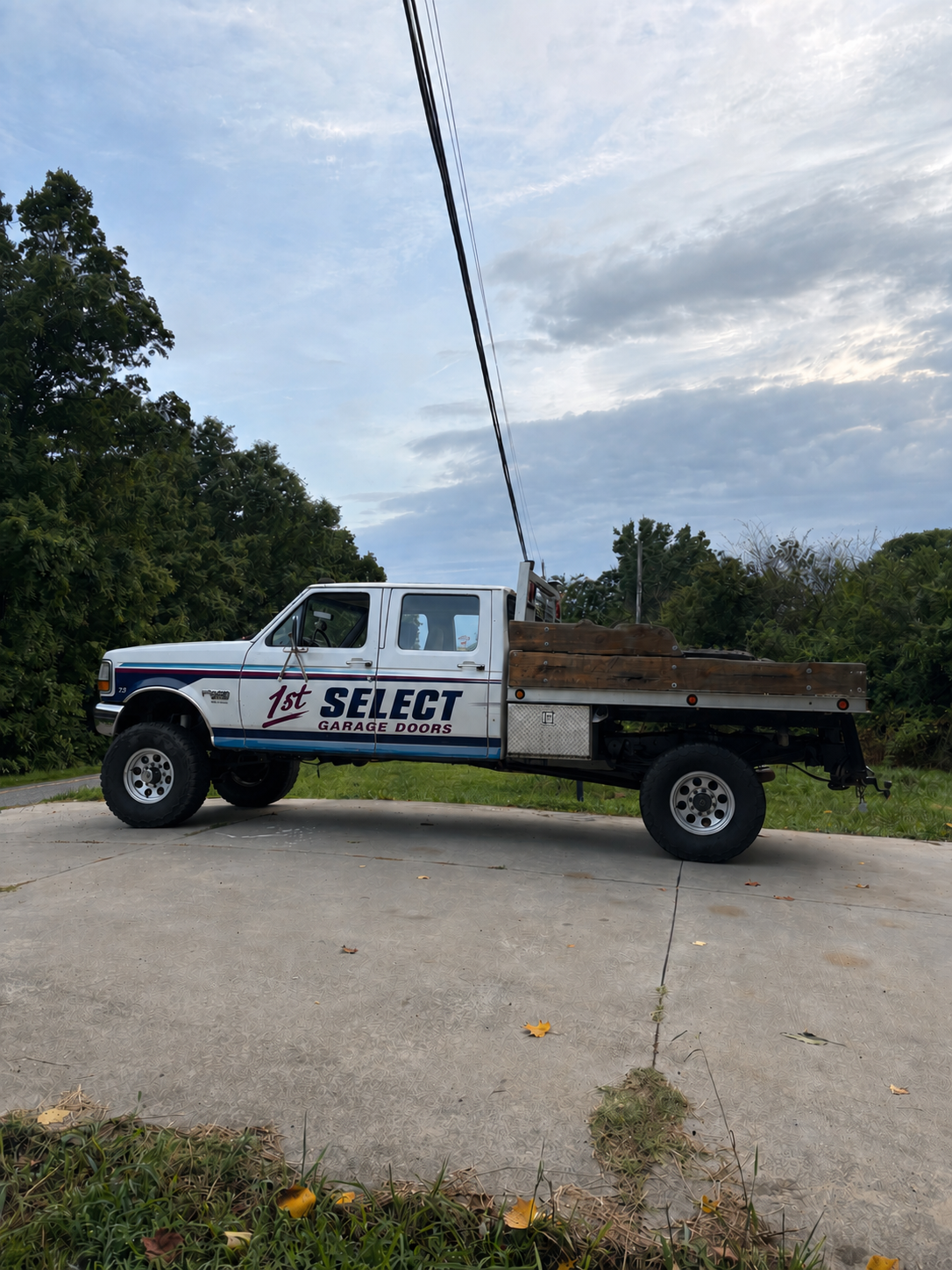 A white pickup truck with a flatbed parked on a concrete surface, surrounded by green trees and a partly cloudy sky.