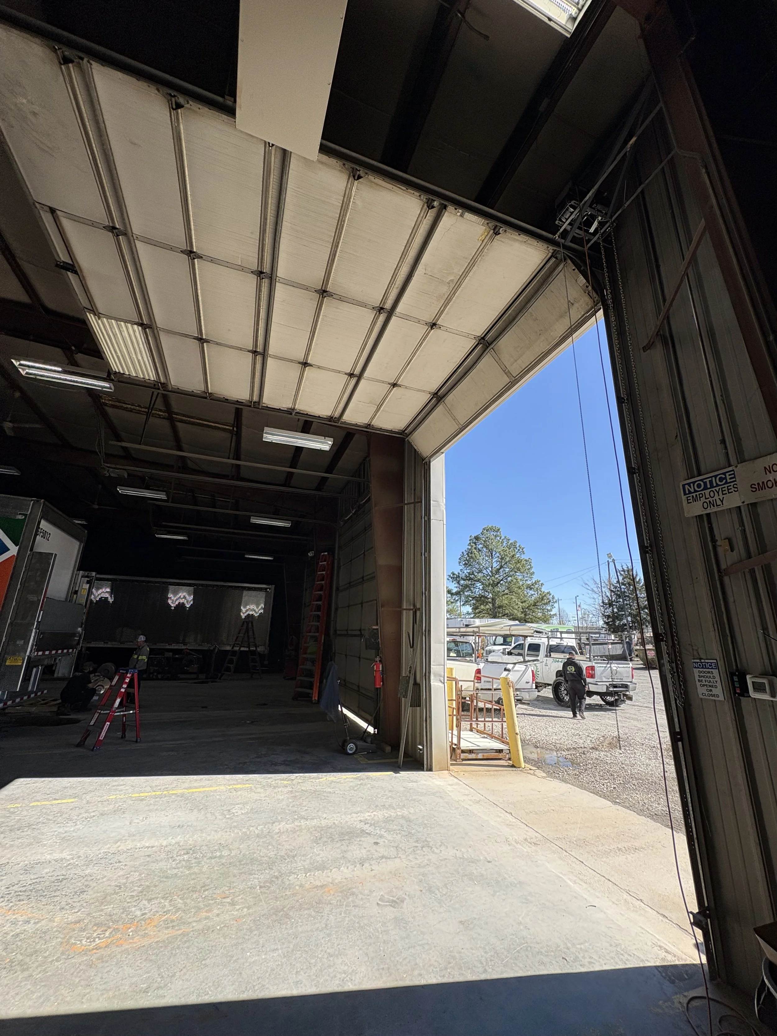 View from inside a warehouse with large open garage door, showing trucks parked outside and workers working inside.
