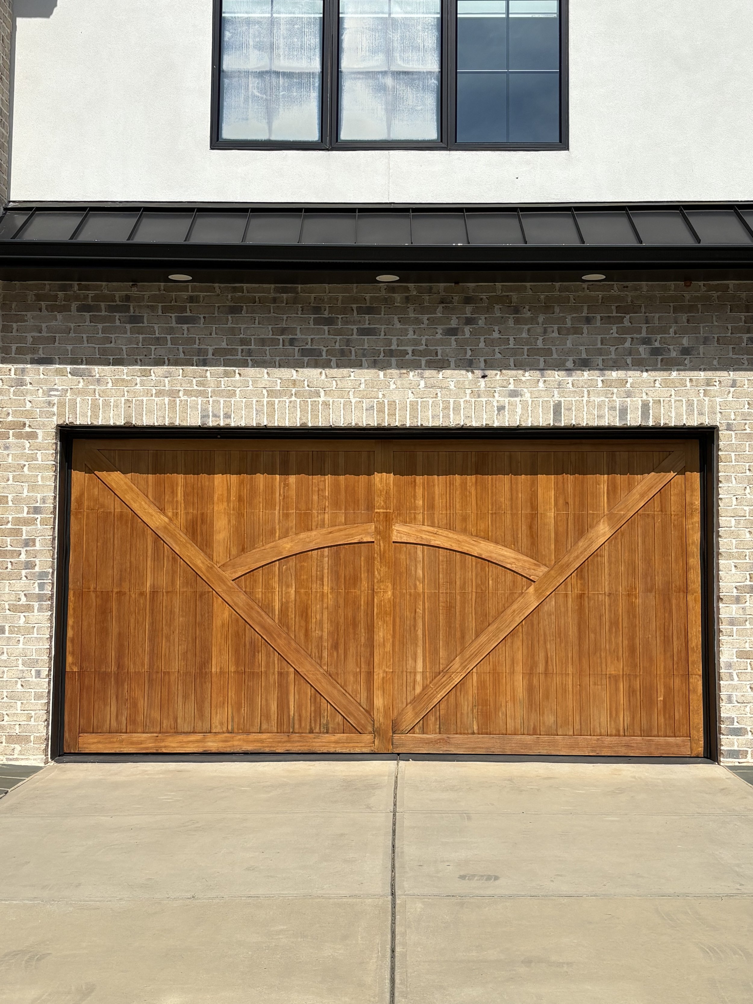 A large wooden garage door with a curved arch design, surrounded by a brick wall and modern upper windows with black frames.