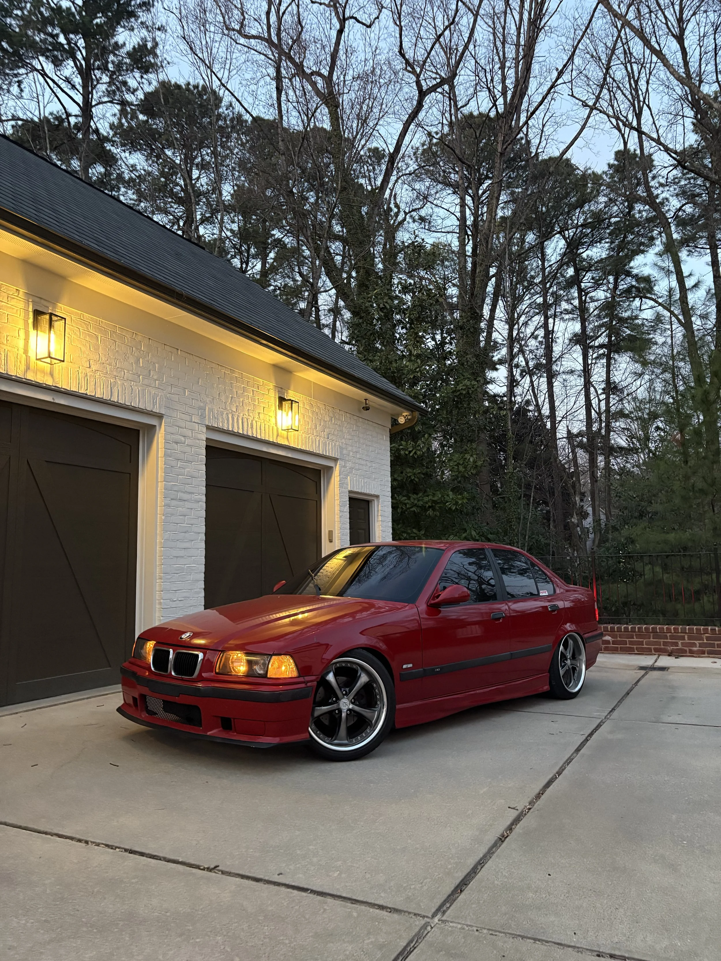 A red BMW car parked on a driveway in front of a white brick house with a dark garage door and outdoor lights. Behind the house, there are tall trees with bare branches, indicating a winter or early spring setting, during twilight or early evening.