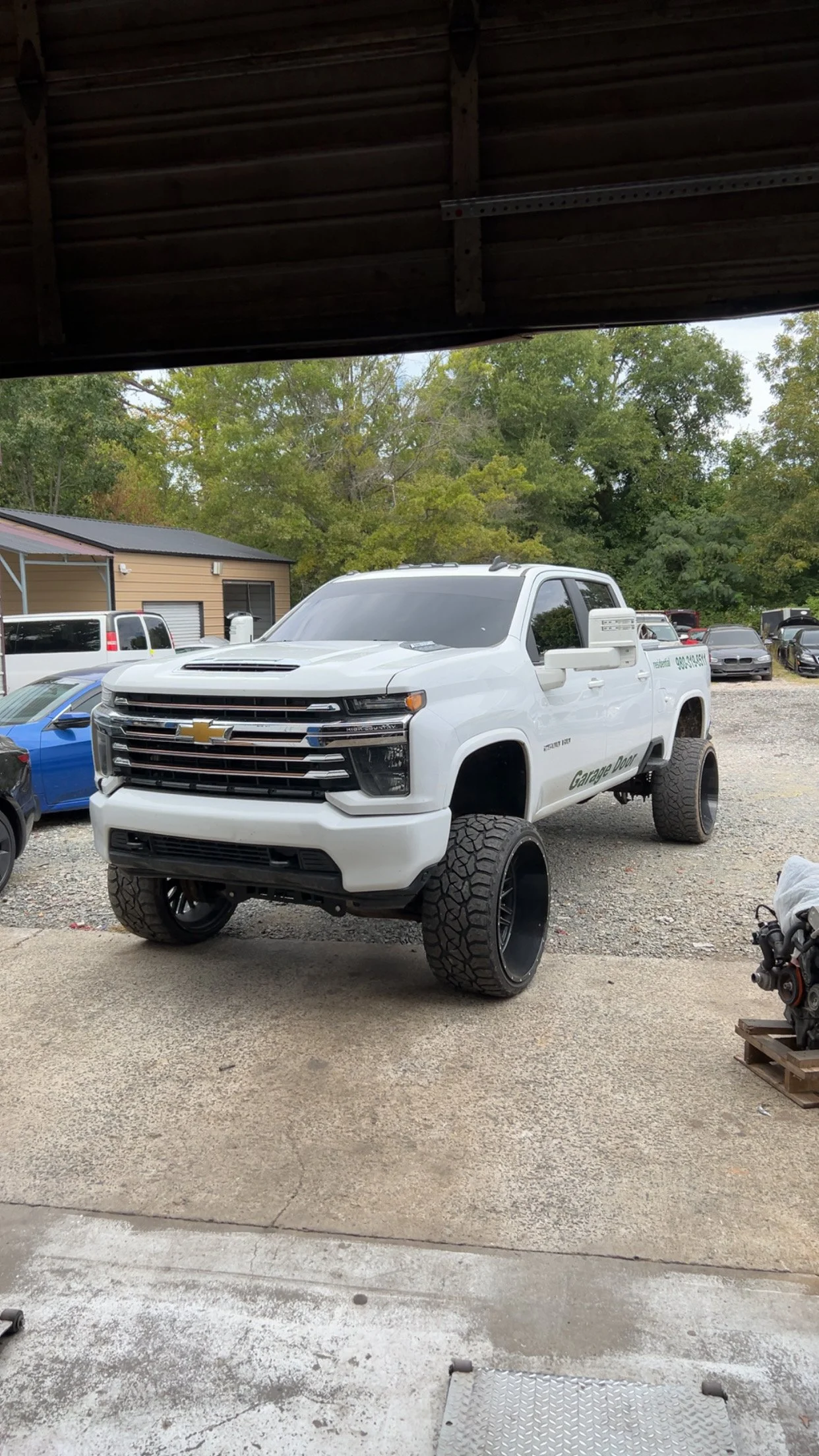 A white lifted Chevrolet truck with large off-road tires parked outdoors near a garage and other vehicles, with trees in the background.