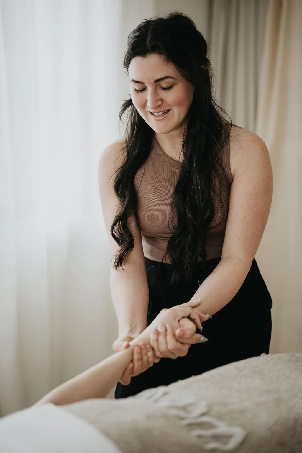 A woman with long dark hair smiling while holding hands of someone lying on a bed.