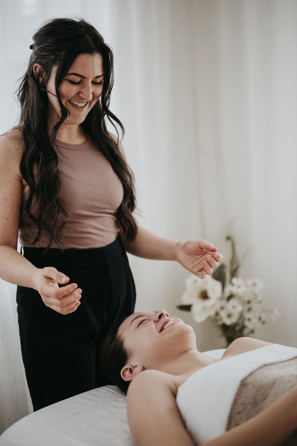 A woman with long dark hair smiling at a person lying on a massage table, relaxing during a massage session.