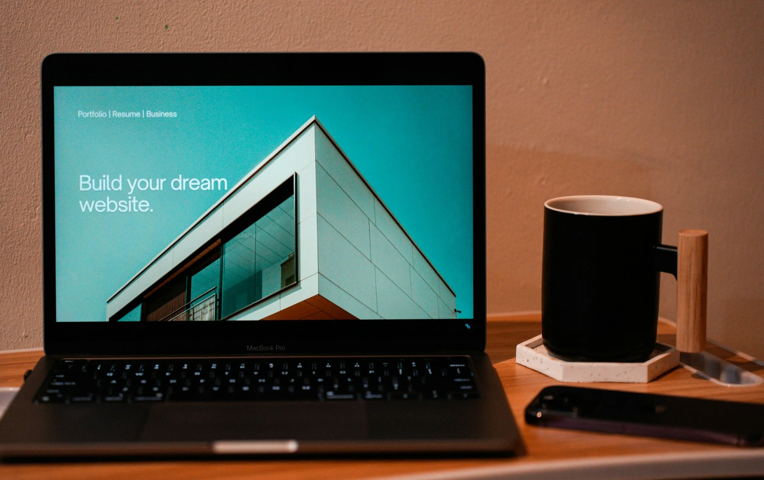 Close-up of a laptop on a wooden desk, with a black mug and coaster to the right, and a smartphone partially visible at the bottom. The laptop screen displays a website with a modern architectural building and the text 'Build your dream website.'