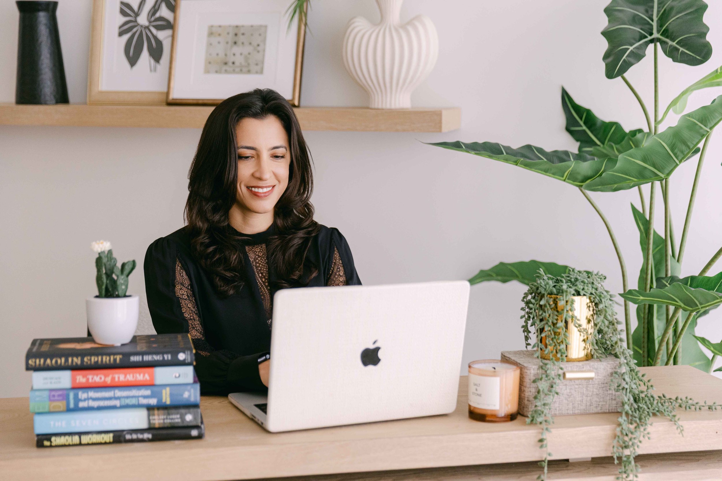 A woman with dark wavy hair smiling while working on a laptop at a desk surrounded by potted plants and stacked books.