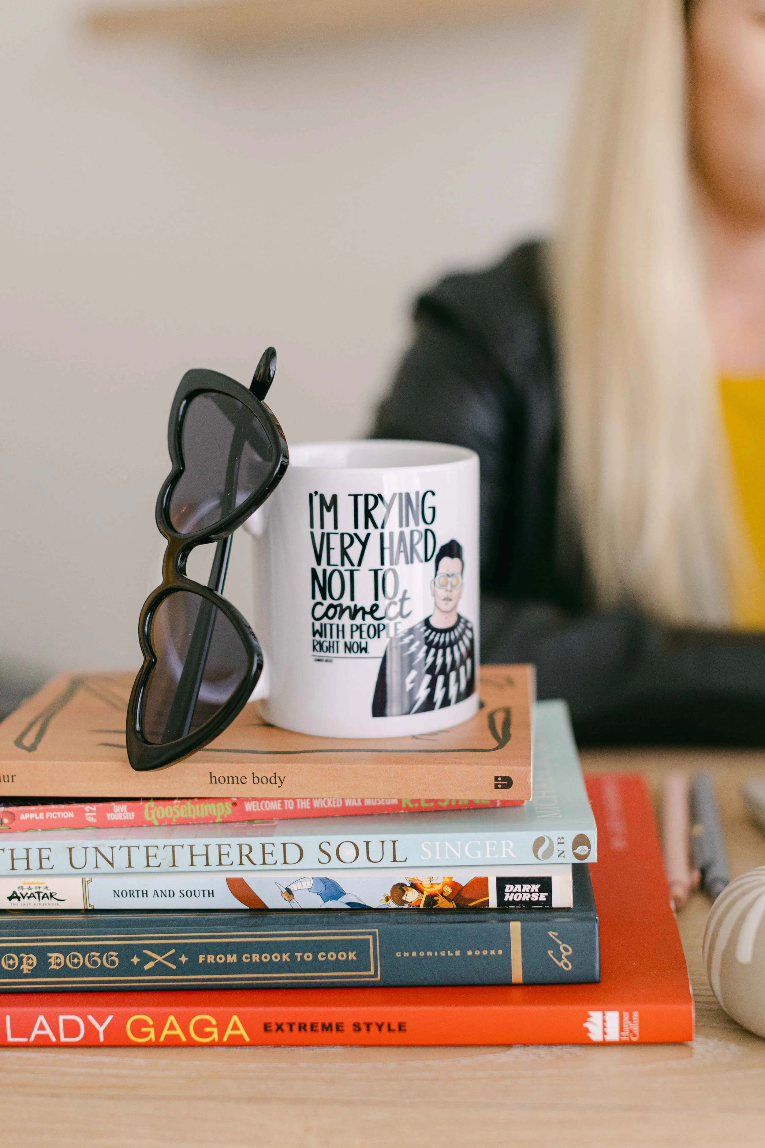A stack of books on a table with a mug and sunglasses on top. The mug has text that says "I'm trying very hard not to connect with people right now" and an illustration of a woman. The books include titles like "The Untethered Soul," "Dark Horse," and "Lady Gaga: Extreme Style."