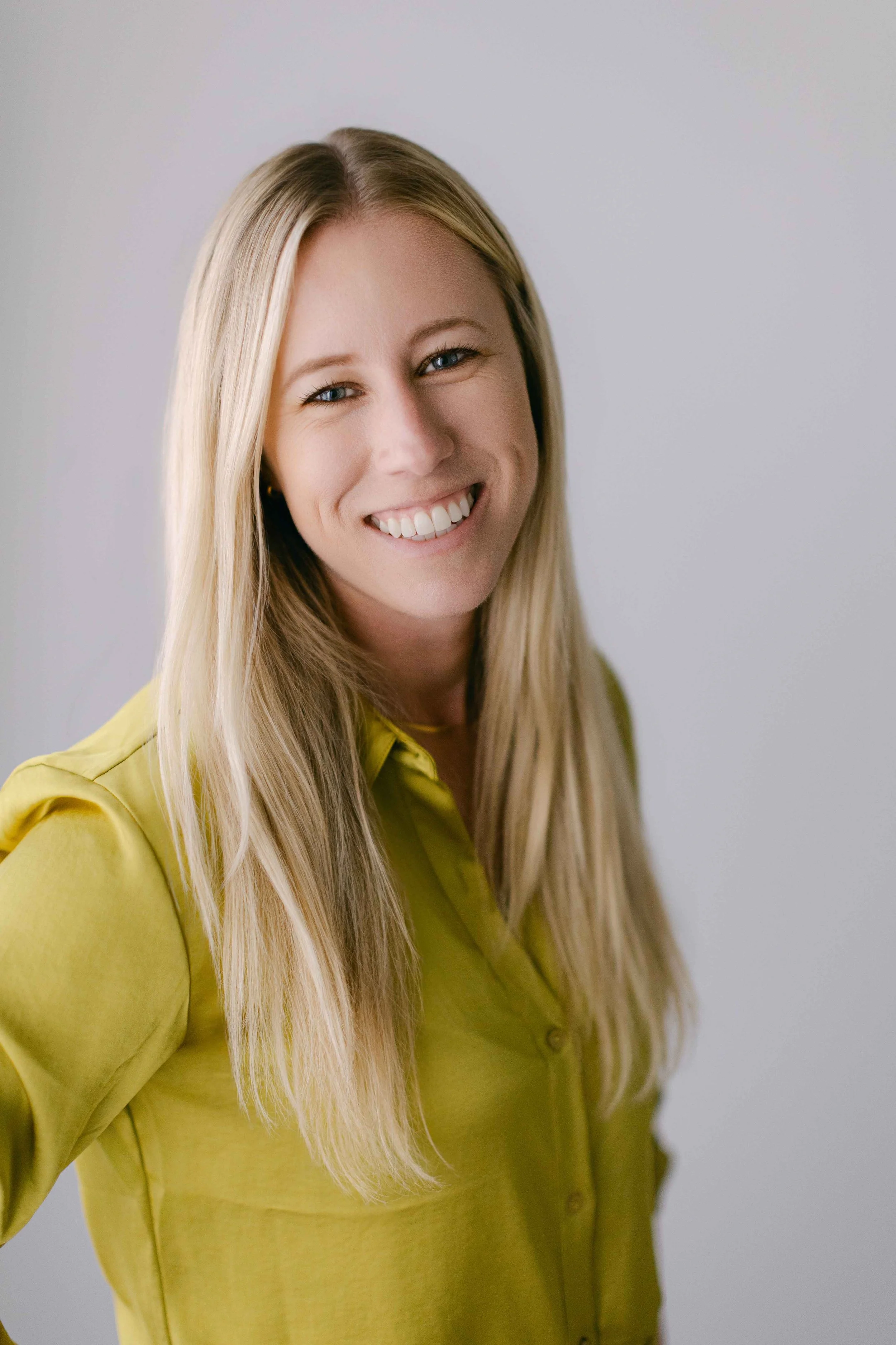 Portrait of a smiling woman with long blonde hair, wearing a yellow shirt, against a plain light background.