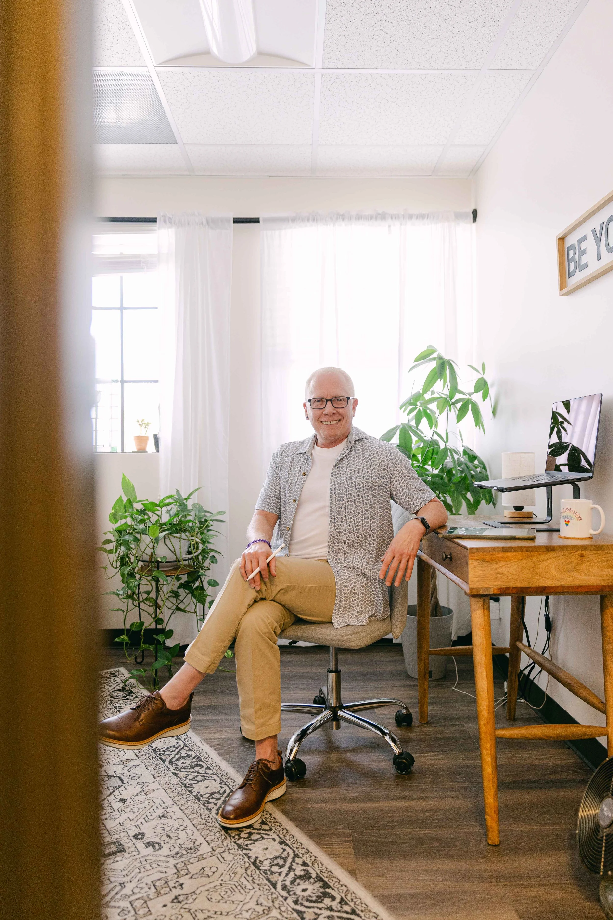 A smiling man with glasses, white hair, and casual clothes sitting in an office, surrounded by plants, with a window and computer on a desk behind him.