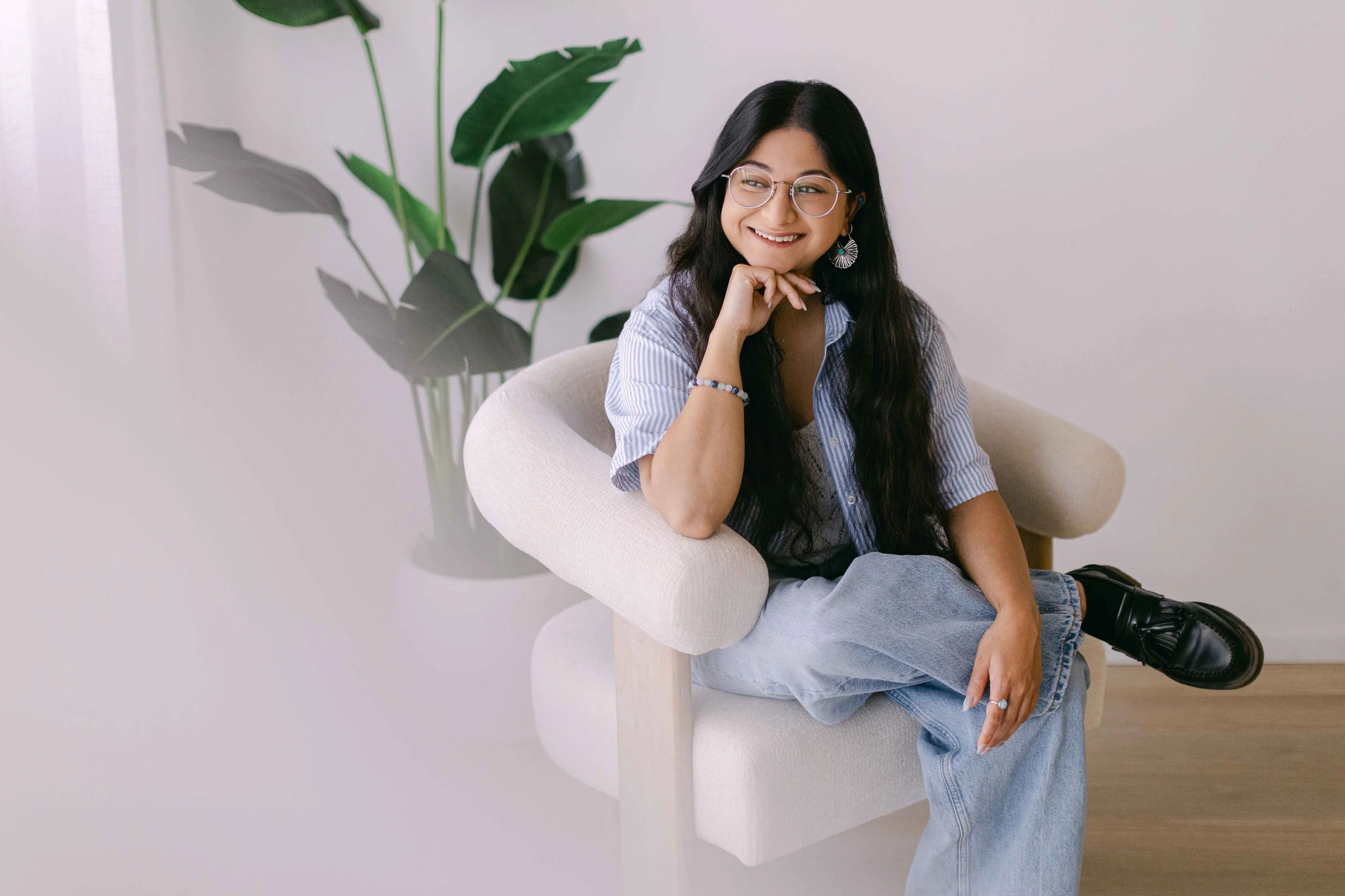 A woman with long dark hair, wearing glasses, a striped shirt, and jeans, sits on a beige chair with her legs crossed, smiling and resting her chin on her hand. A large green plant is in the background against a plain white wall.