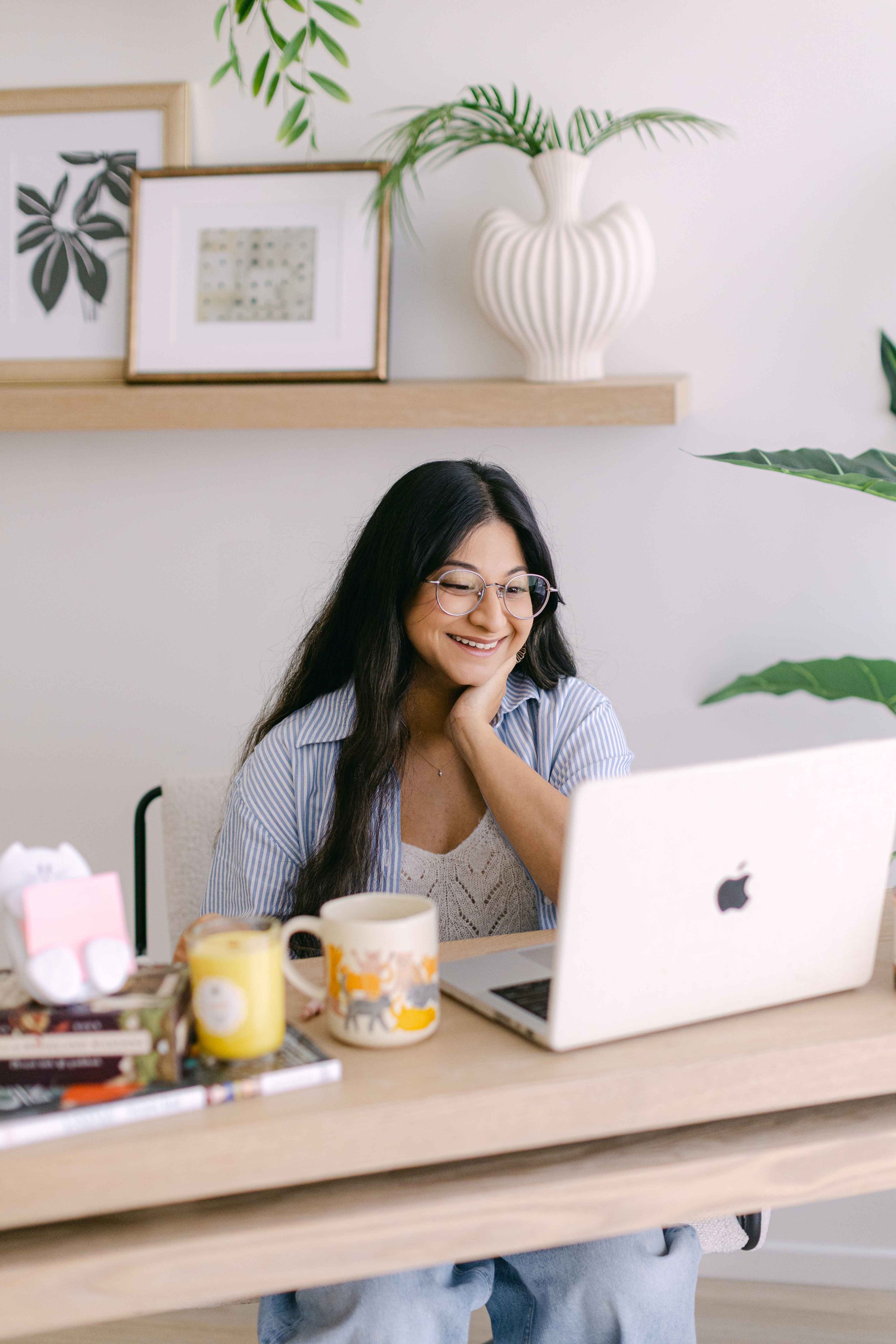 A woman with long dark hair, glasses, and a striped shirt sitting at a desk, smiling while looking at a laptop, with a white coffee mug and snacks on the desk. Behind her, there are framed art prints and a white vase with green plants on a wooden shelf.