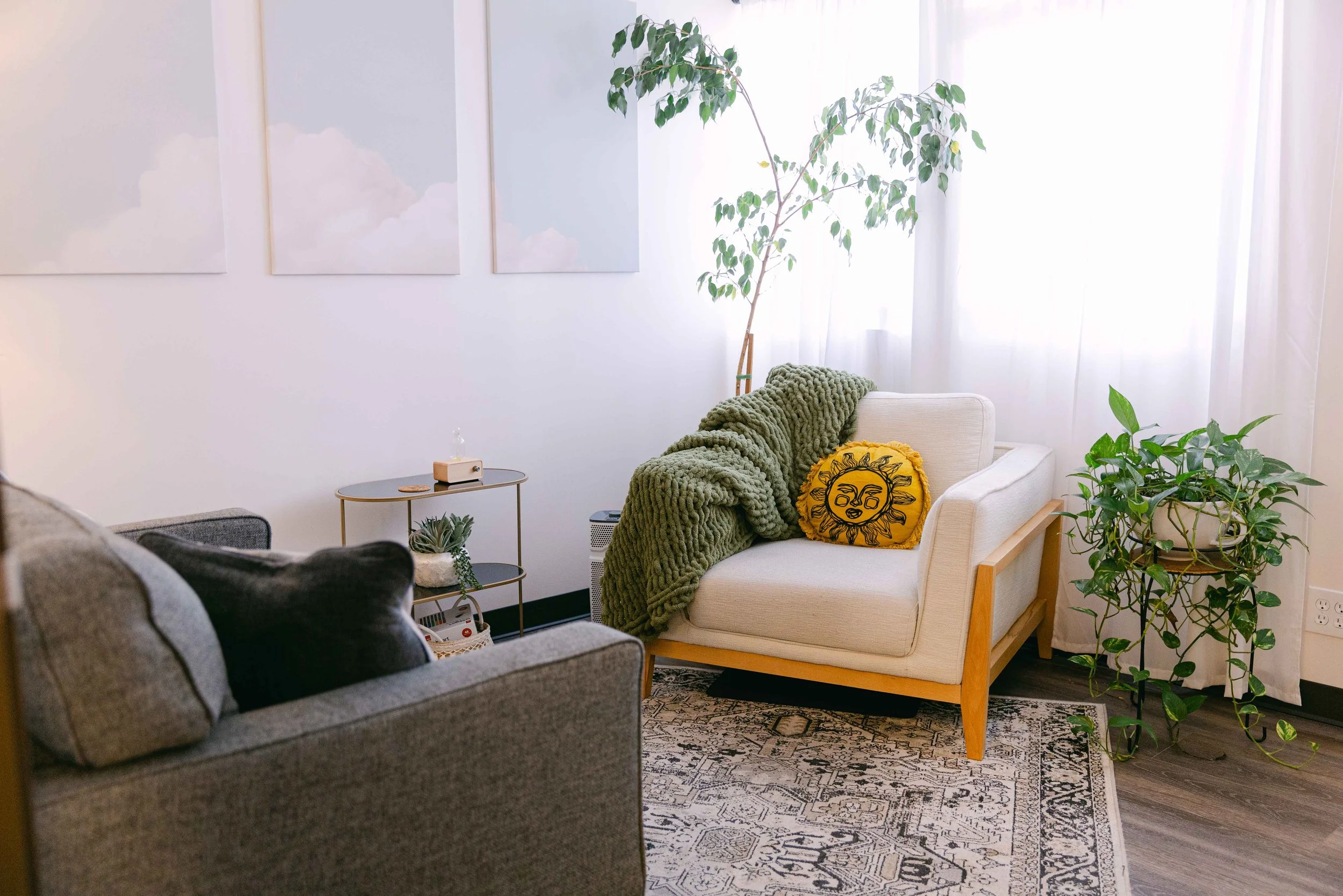 Living room with two sofas, one gray with cushions and one cream with a green chunky blanket and a yellow sun face pillow, wooden framed armchair, potted plants, sunlit white curtains, three pastel cloud paintings on the wall, small round side table with plants and a tissue box, patterned rug, hardwood floor.