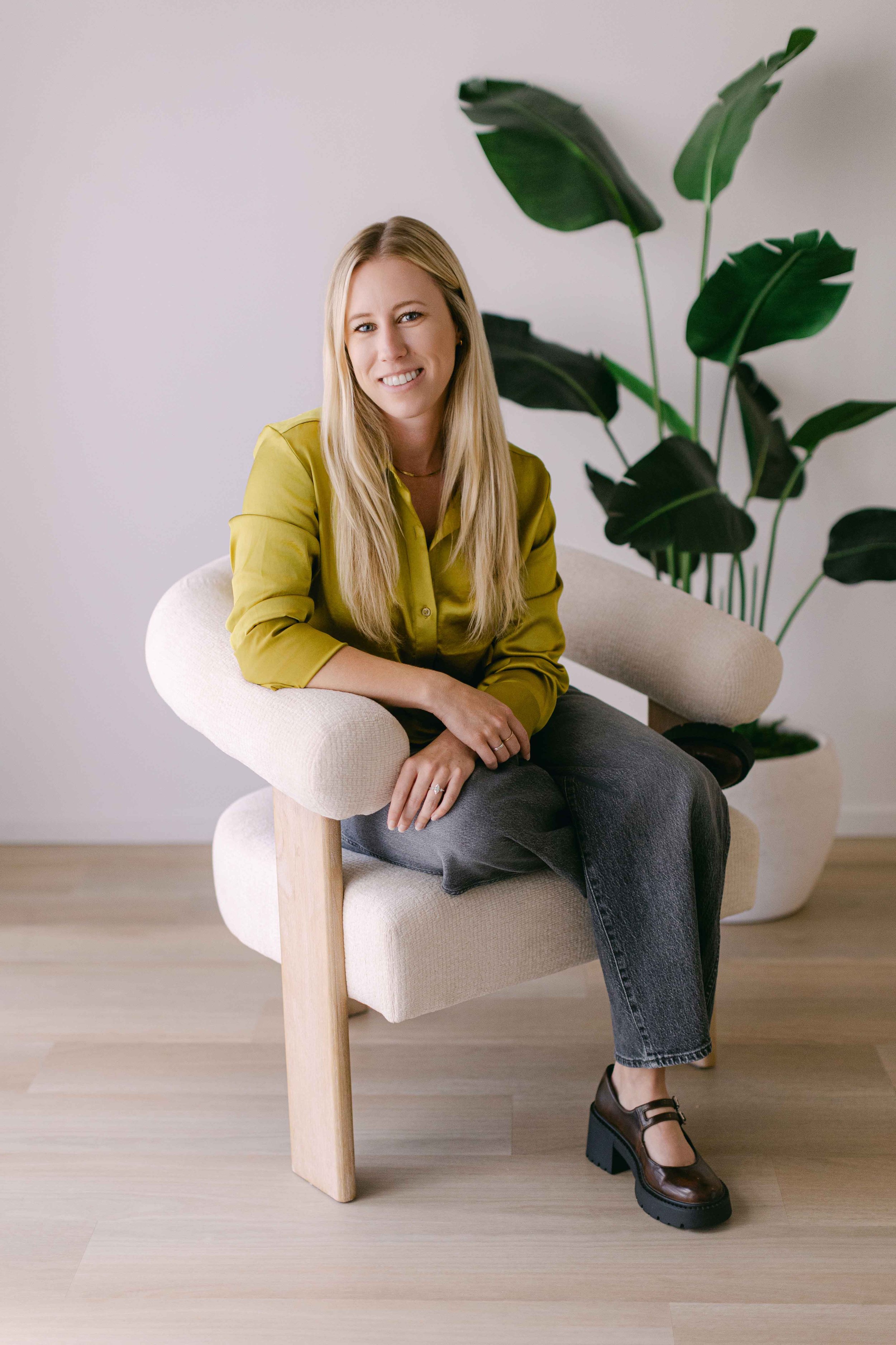 A woman with long blonde hair smiling while sitting on a beige armchair with wooden legs. She is wearing a yellow shirt, dark jeans, and brown shoes, with a large green leafy plant in the background, against a plain light-colored wall.