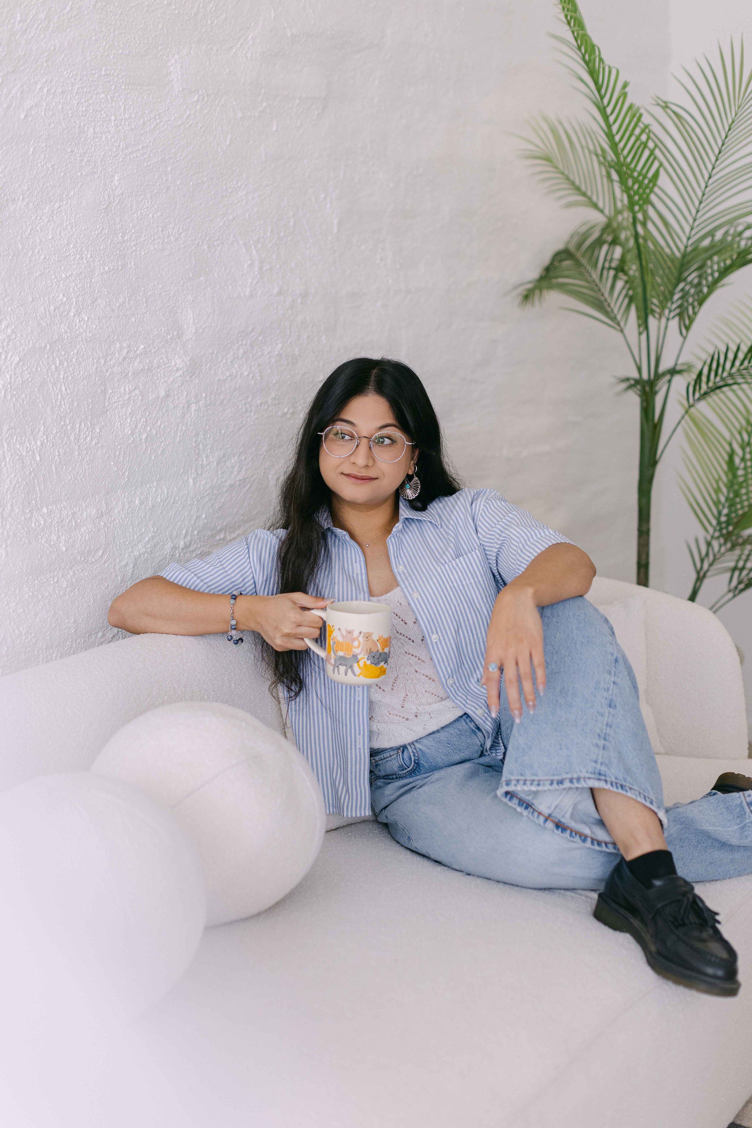 A young woman with long black hair, glasses, and earrings, sitting on a white sofa, holding a colorful mug, wearing a blue striped shirt and ripped jeans, with a large green plant behind her, in a bright room with a textured white wall.