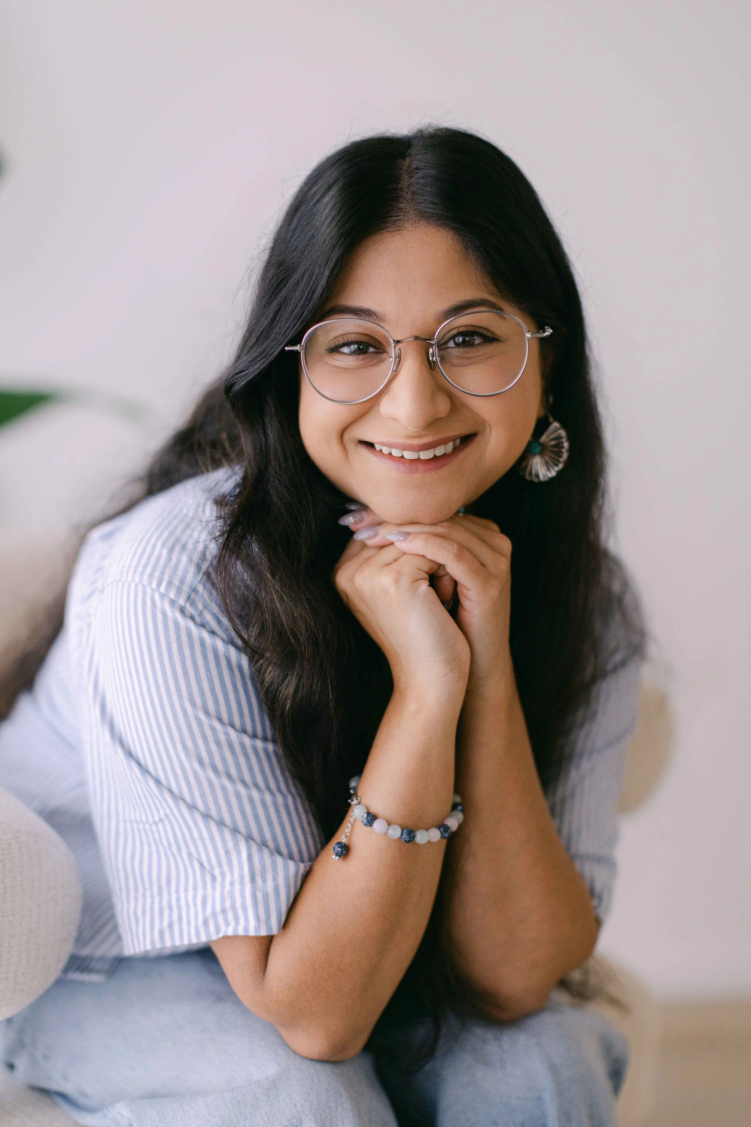 Close-up portrait of a young woman with long dark hair, wearing glasses, smiling with hands under chin, sitting on a light-colored couch, wearing a striped shirt and jewelry.