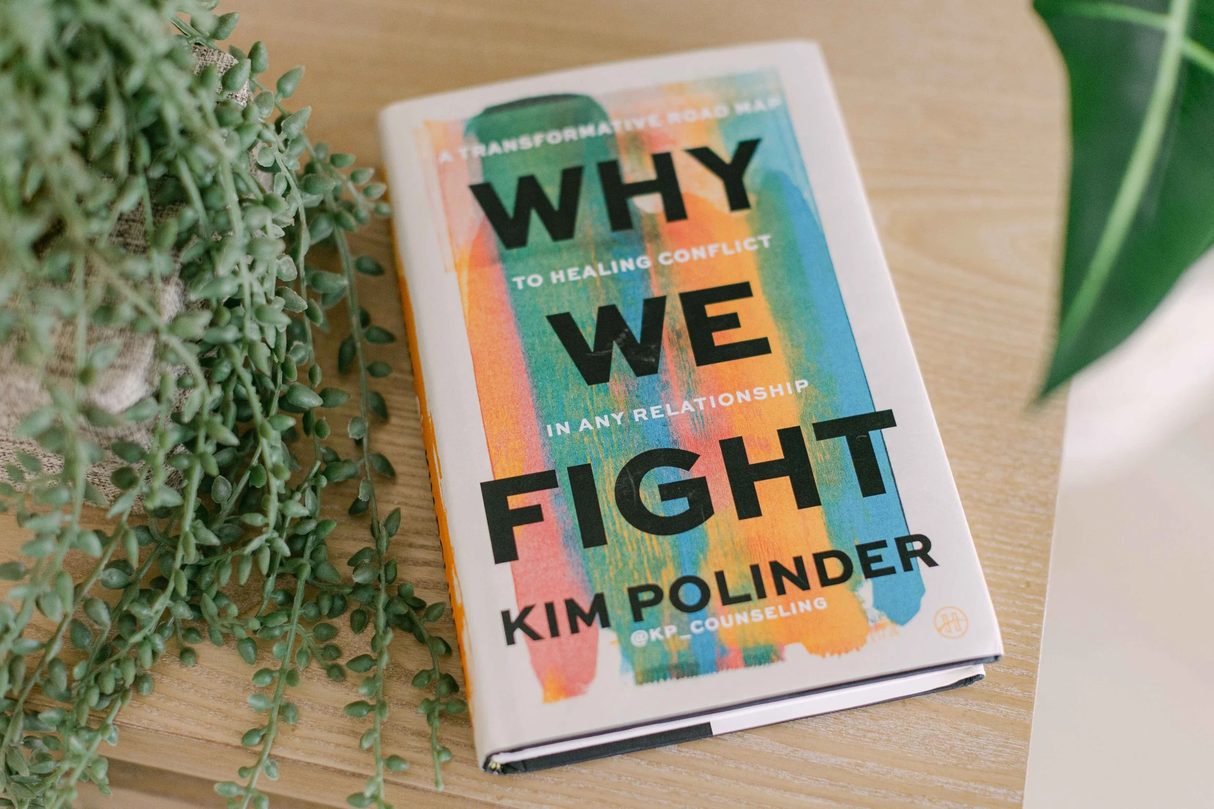 A colorful book titled 'WHY' by Kim Polinder is lying on a wooden surface, with a green potted plant and another plant with broad leaves nearby.