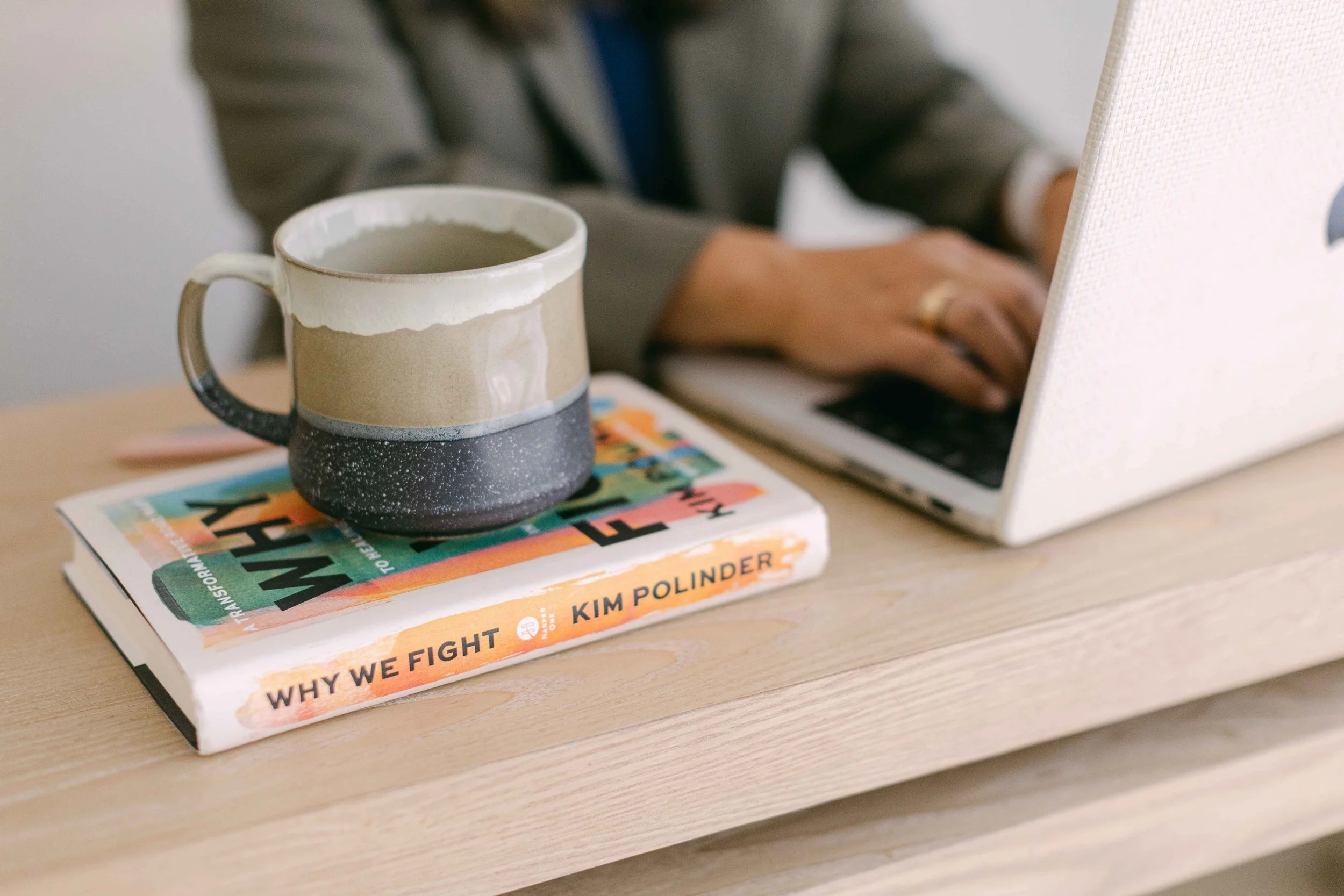A beige, black, and white speckled coffee mug filled with coffee, resting on top of a stack of three books on a light wooden surface, with a person working on a laptop in the background.