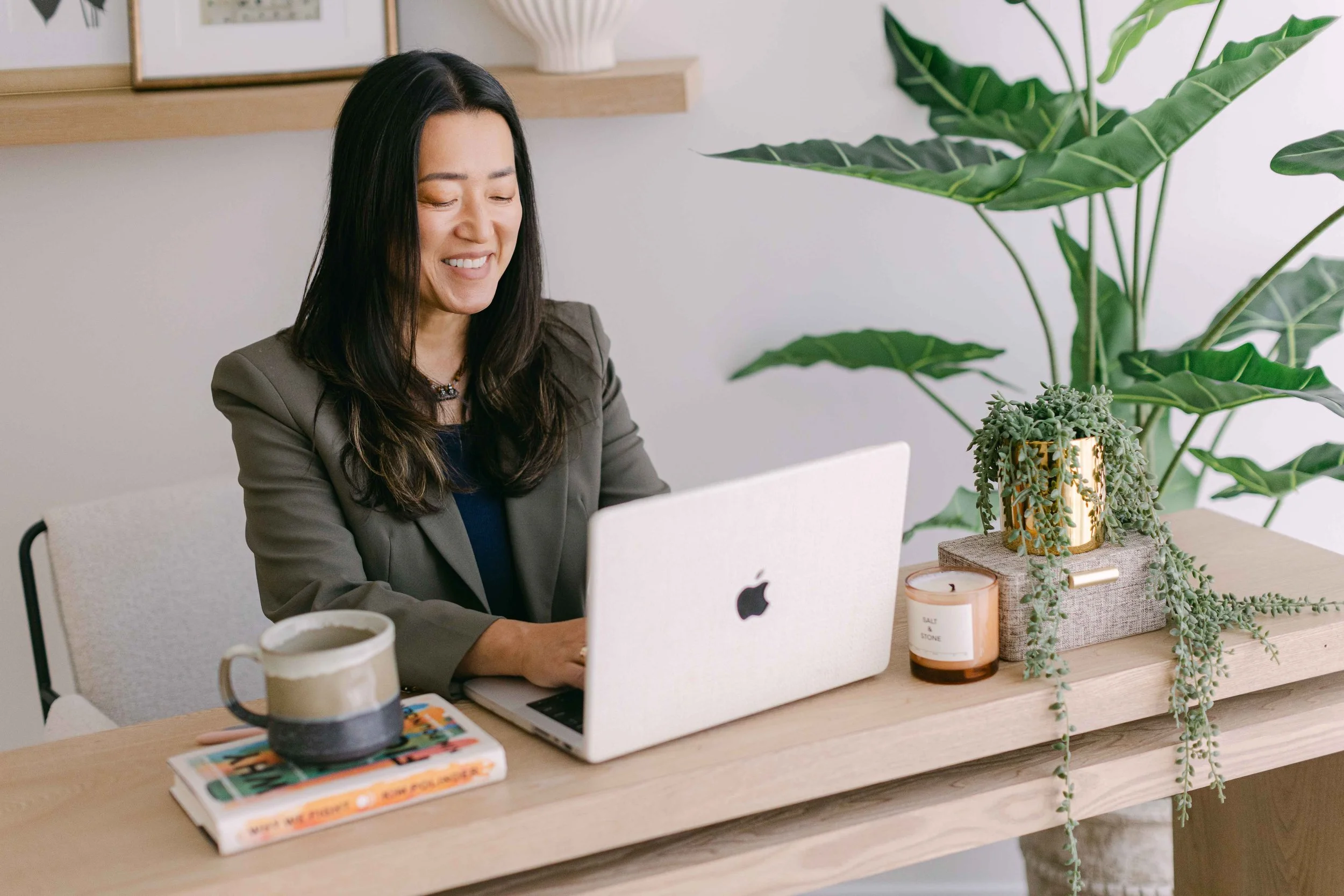A woman sitting at a light wood desk working on a MacBook computer, with a large green houseplant and a smaller potted plant on the desk, and a cup of coffee nearby.