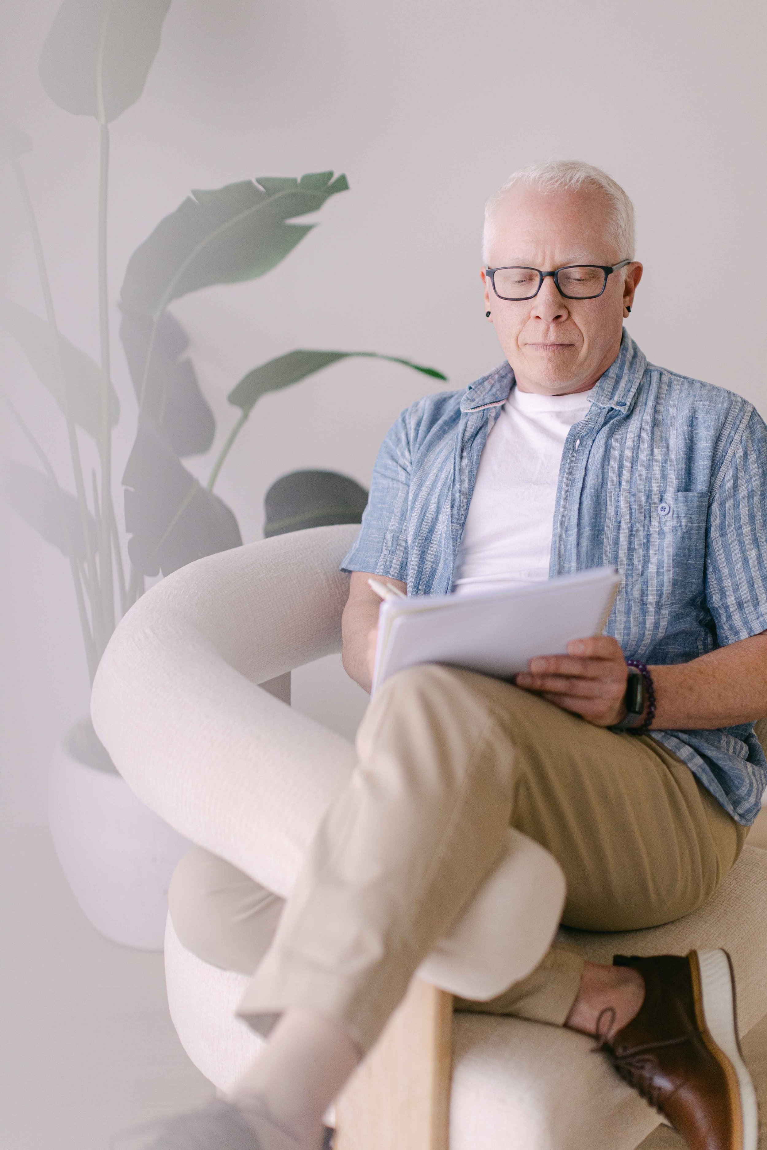 An older man with white hair and glasses is sitting comfortably on a modern white chair, reading a book or magazine. He is dressed casually in a blue plaid short-sleeve shirt and beige pants, wearing brown shoes. Behind him, a tall green plant with large leaves is visible, set against a plain light-colored wall.