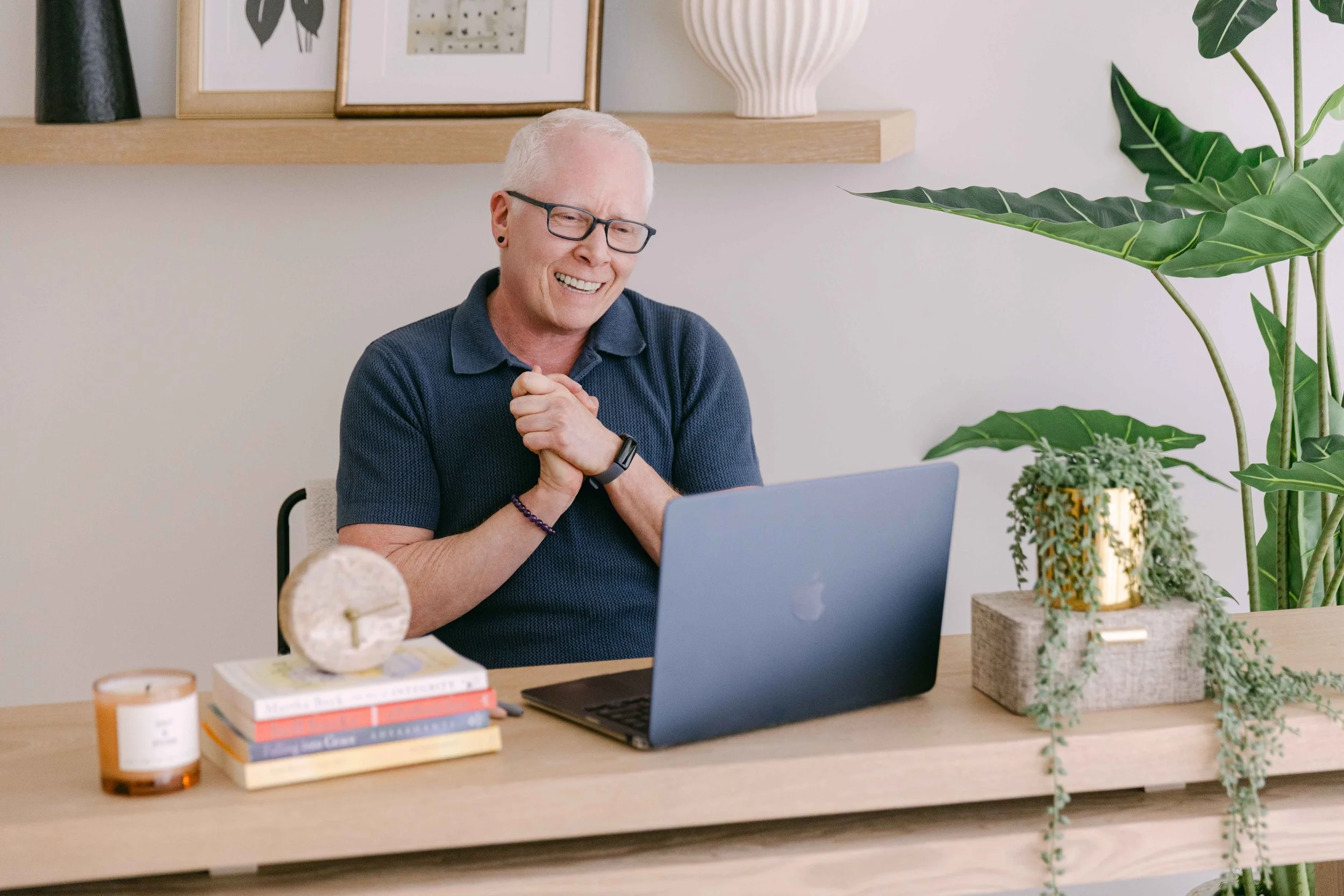 An elderly man with glasses smiling and clasping his hands together in front of a laptop on a wooden desk, surrounded by houseplants, decorative objects, and a small stack of books.