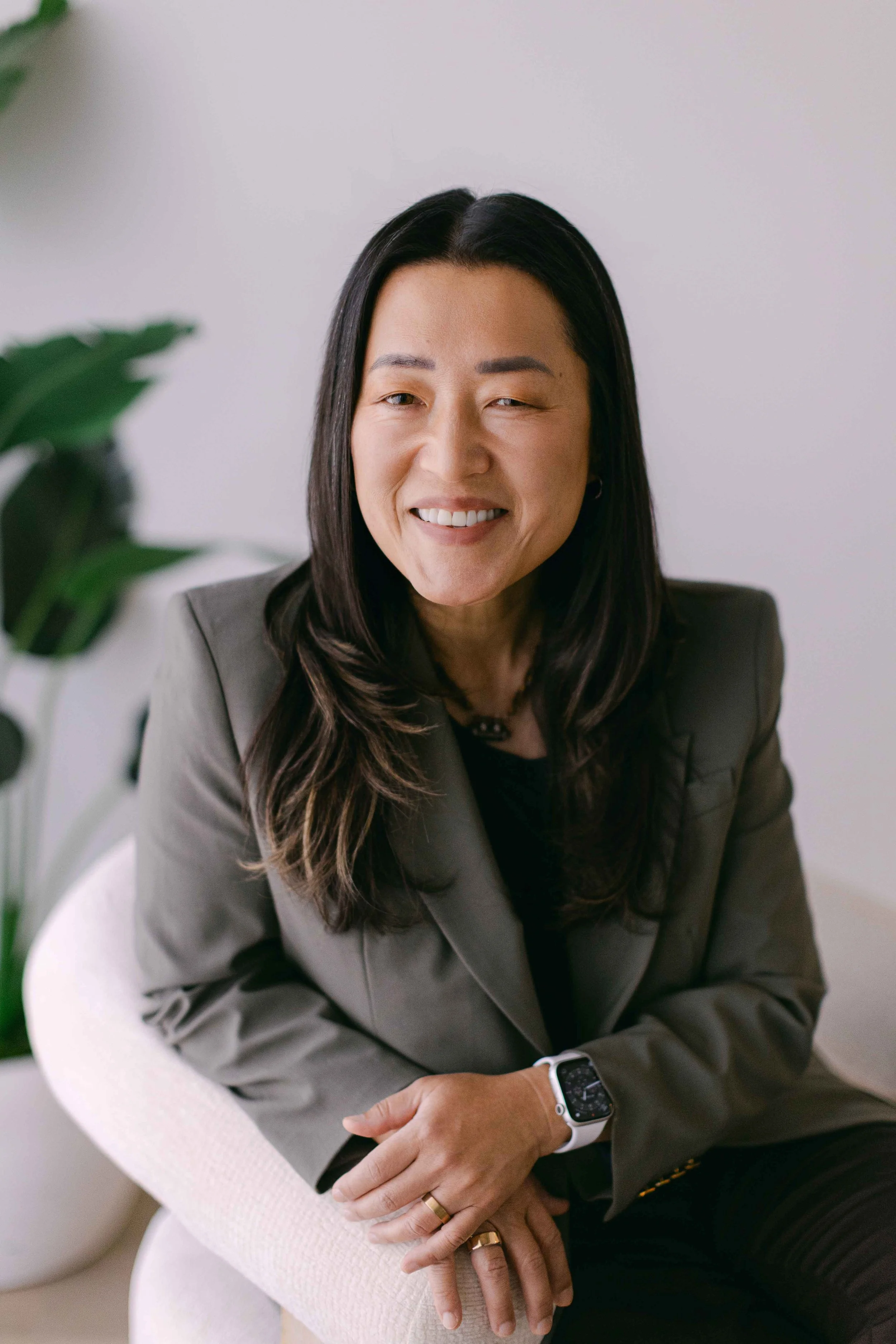 A smiling woman with long dark hair, wearing a gray blazer and a smartwatch, sitting in a white armchair with a blurred green plant in the foreground.