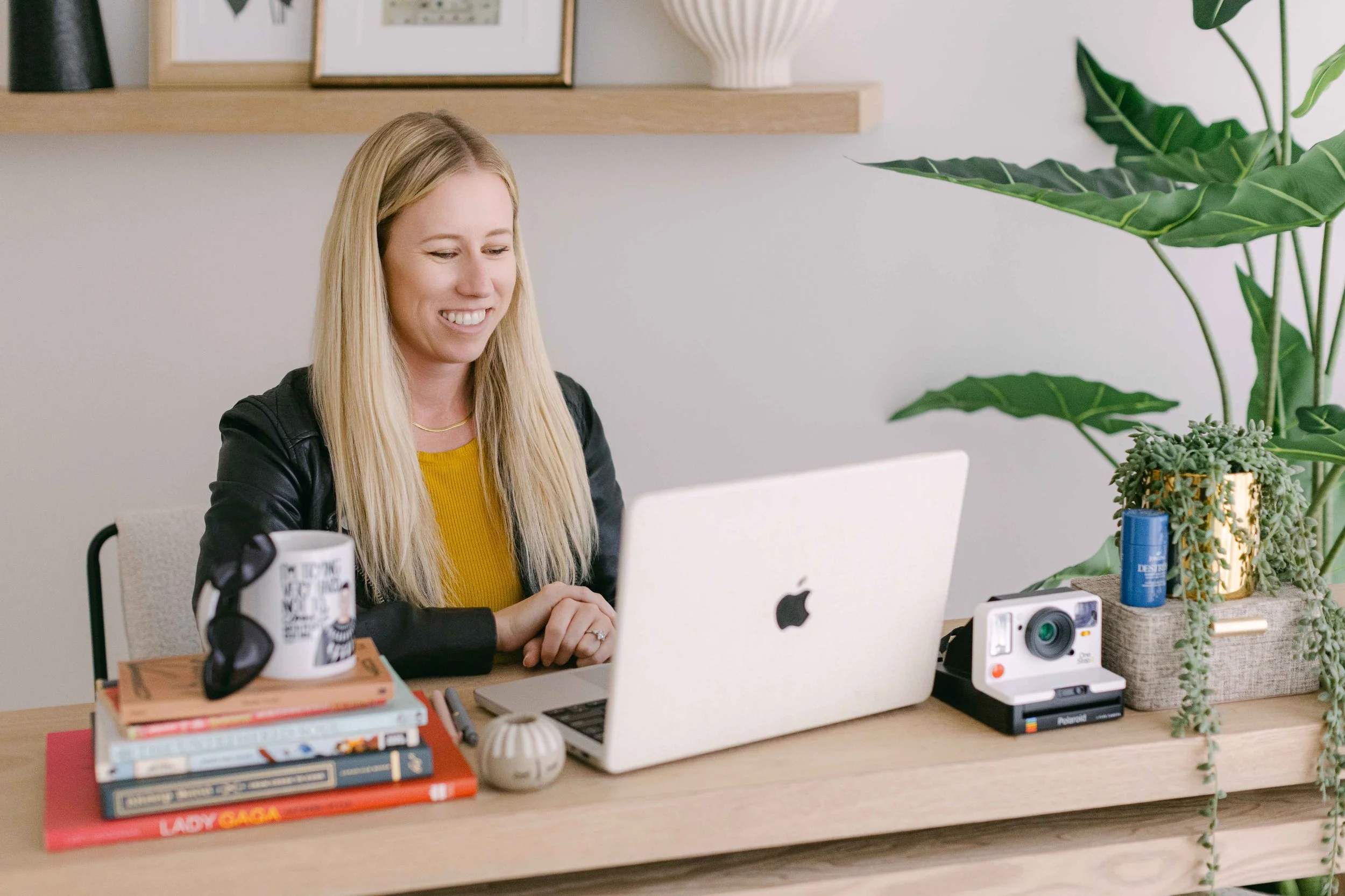 A woman with long blonde hair sitting at a desk with a laptop, surrounded by books, a camera, a mug, and plants in a room with framed pictures on the wall.