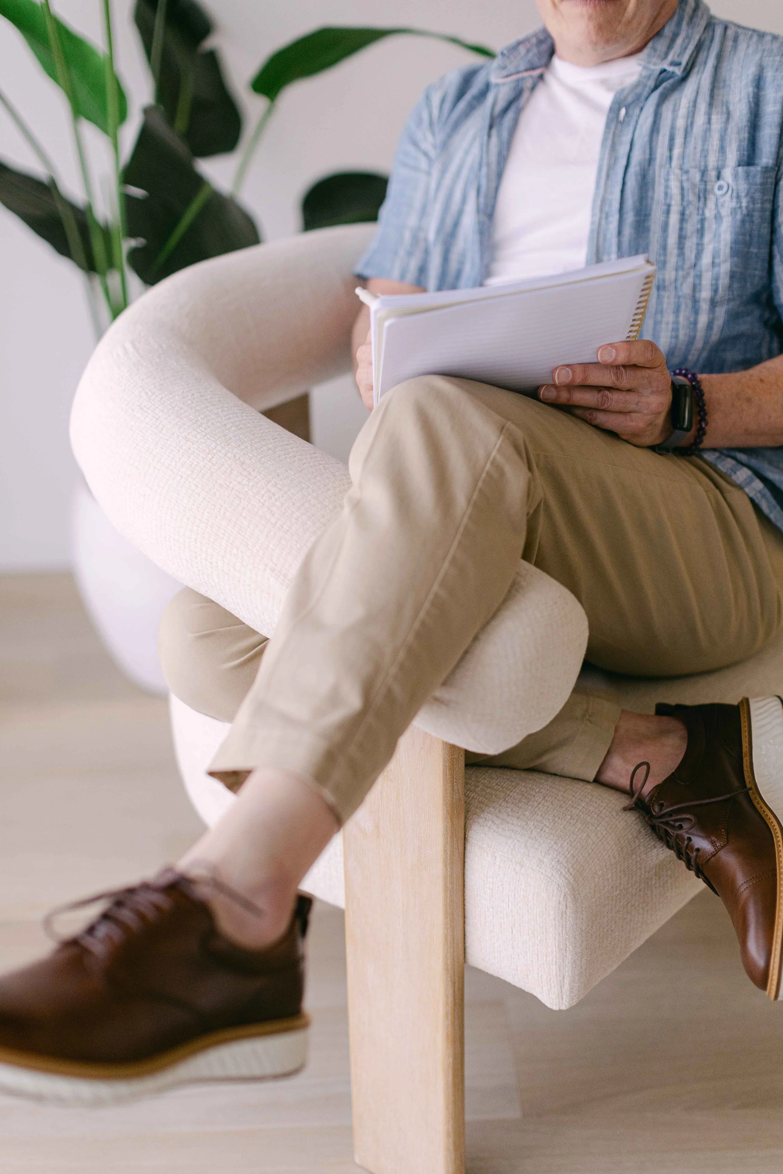 A man sitting on a beige armchair with his legs crossed, wearing beige pants and brown shoes, holding a spiral notebook. There are green indoor plants in the background.