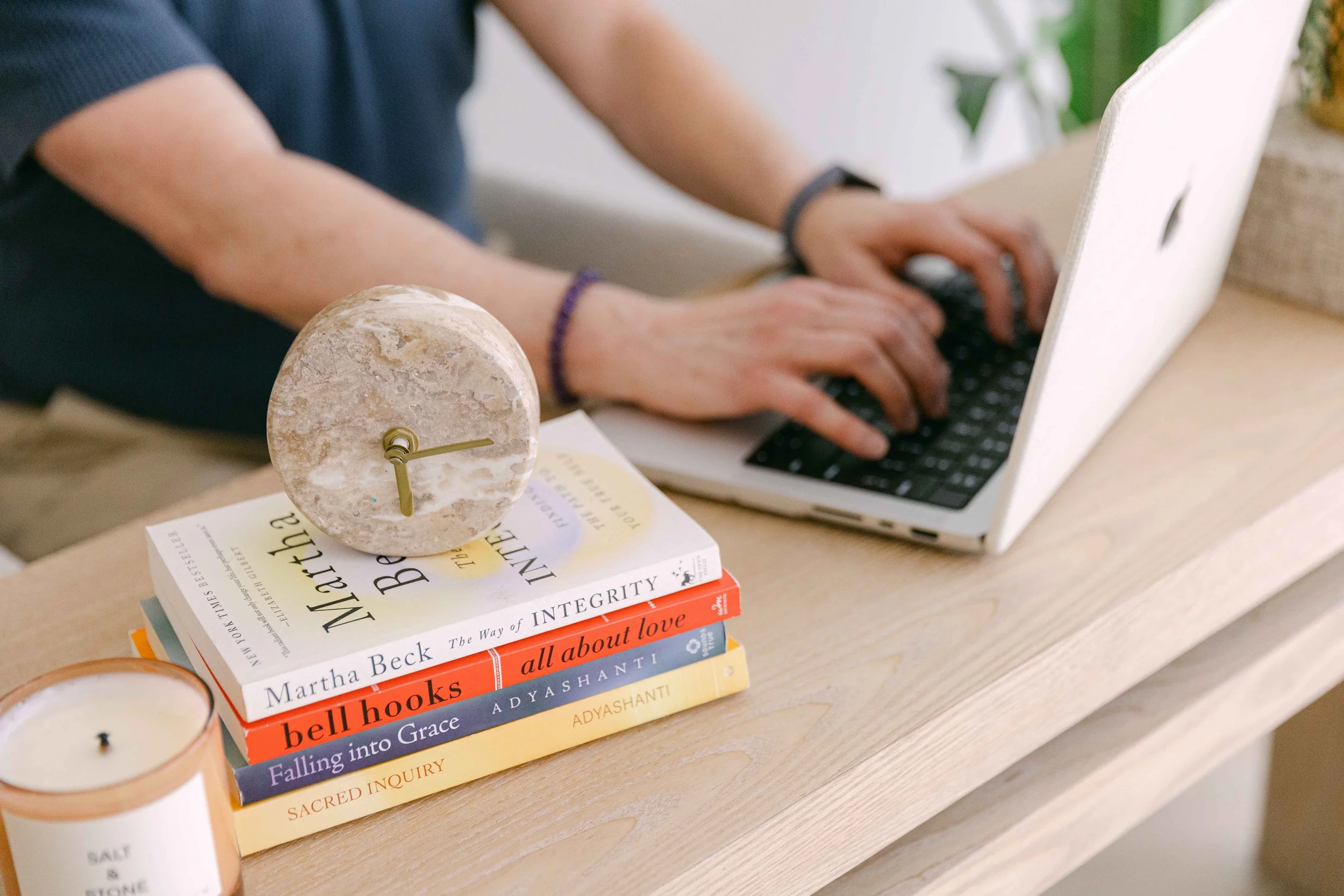 A person typing on a laptop with a stack of books on a table in front of them, a lamp, and a candle nearby.