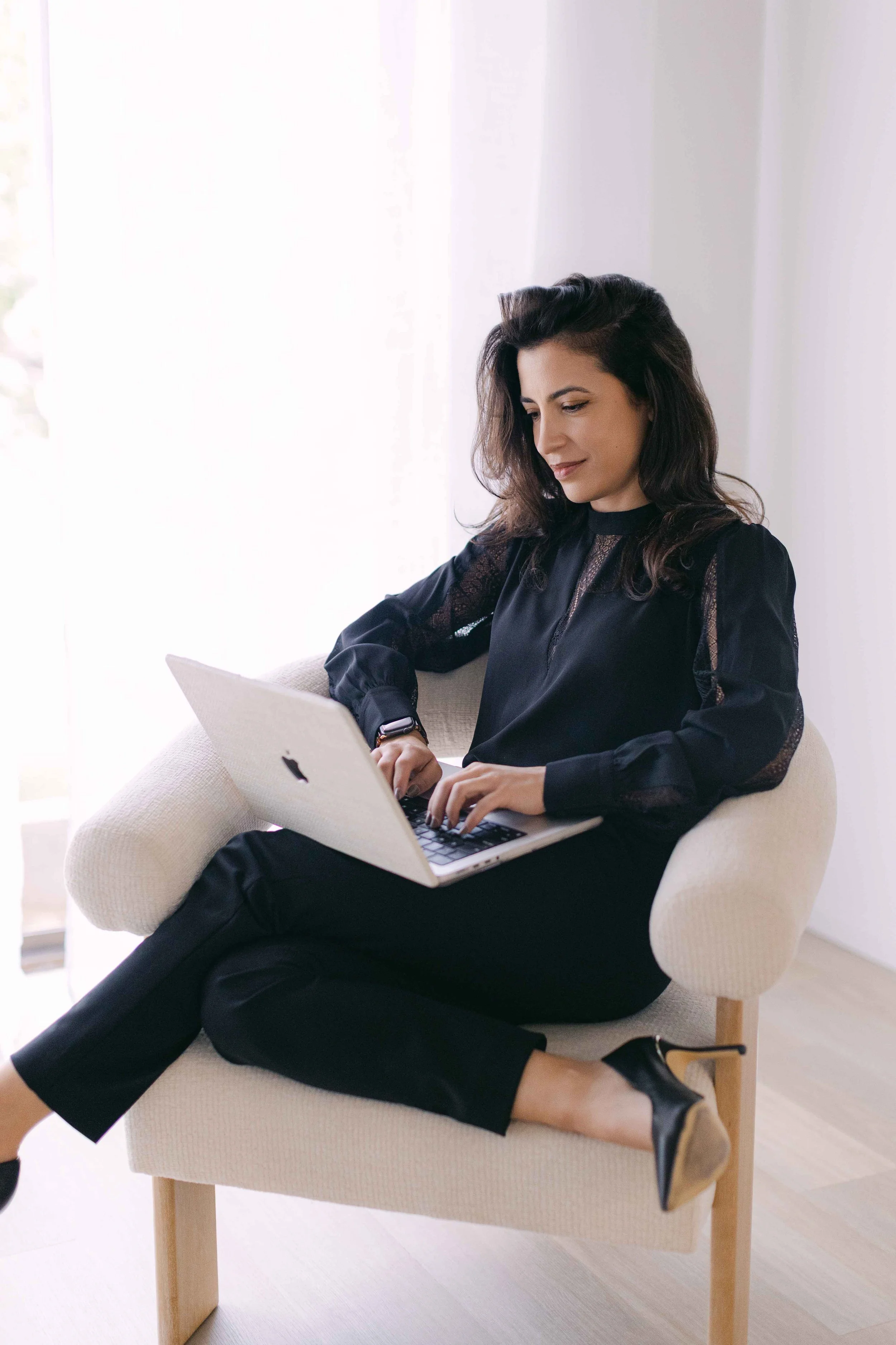 A woman sitting on a beige armchair, working on a laptop with a white exterior and black keyboard, in a room with white walls and a large window with sheer curtains. She is dressed in a black long-sleeve blouse and black pants, wearing black heels, and has dark hair styled in loose waves.