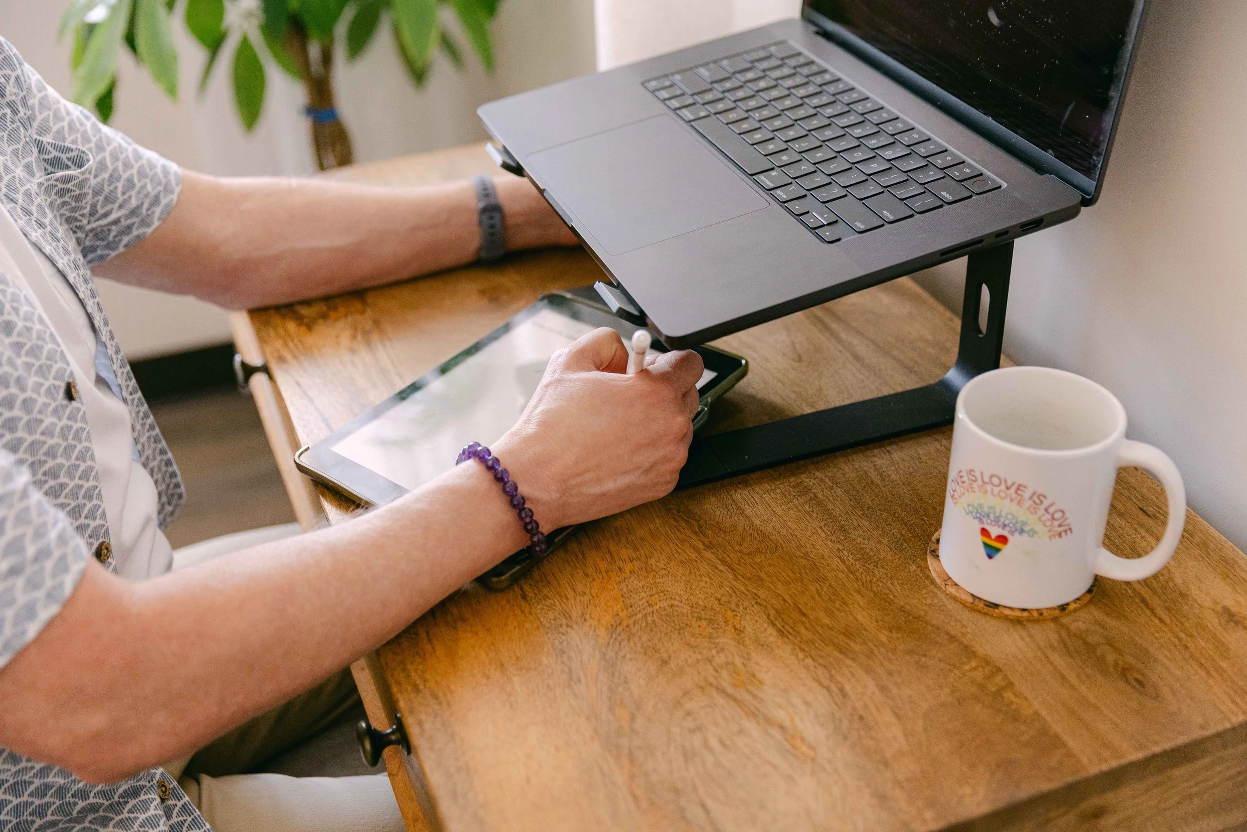 A person with light skin wearing a patterned shirt is sitting at a wooden desk, using a laptop supported by a stand. They are holding a stylus or pen and working on a tablet device. A colorful mug with rainbow and love-themed text is on the desk.