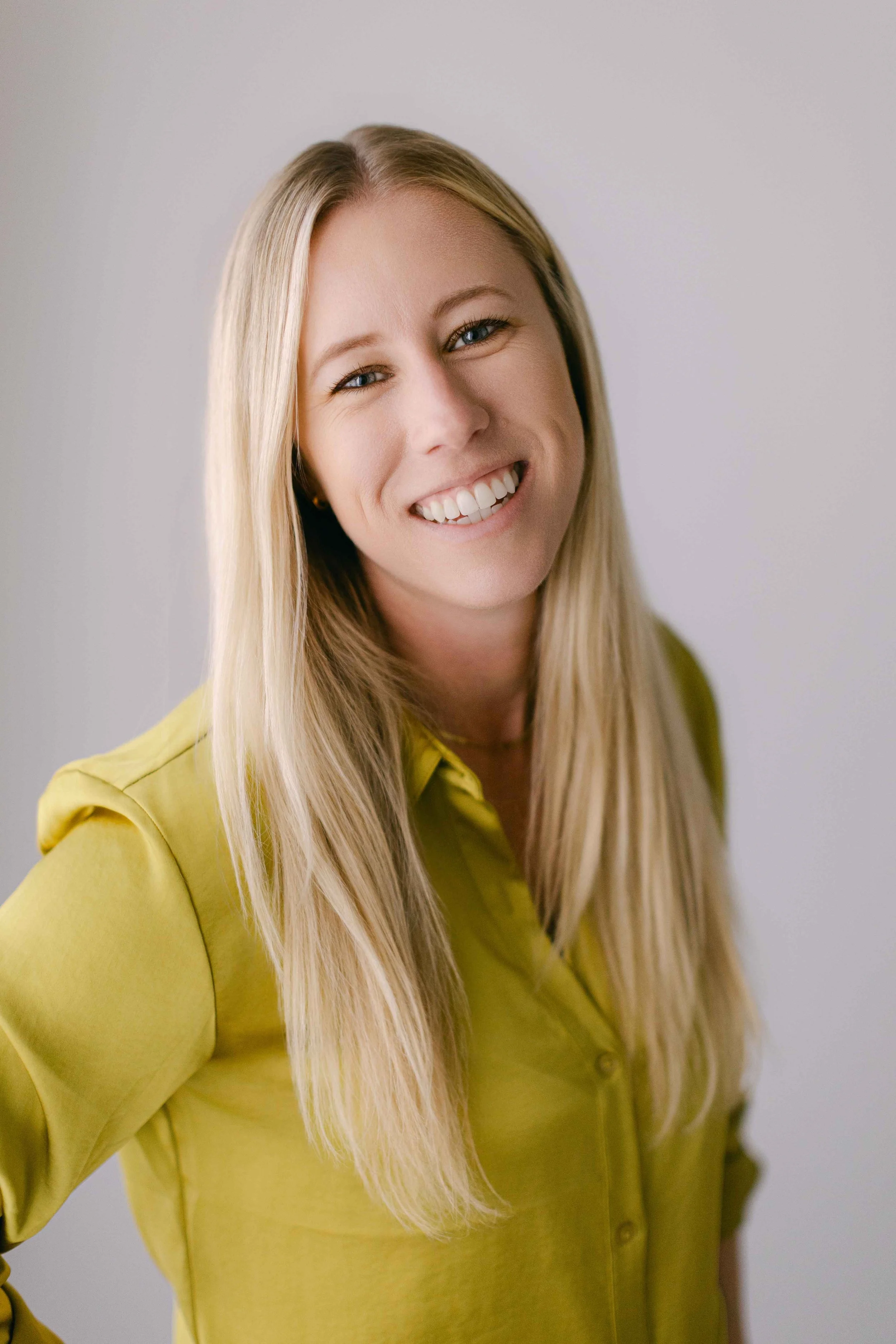 A woman with long blonde hair, blue eyes, and fair skin, smiling, wearing a yellow shirt, standing against a plain light gray background.