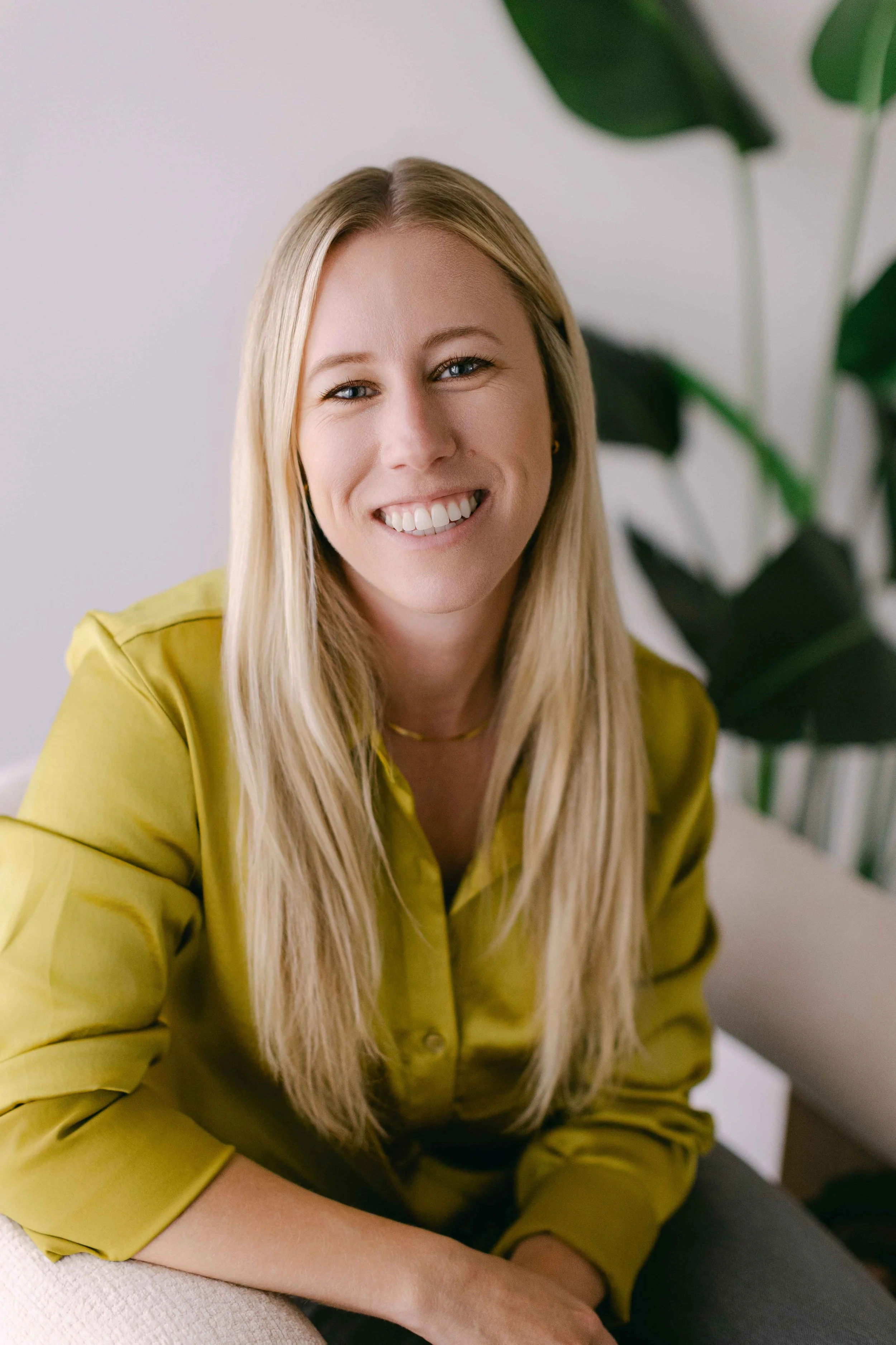 Portrait of a smiling blonde woman wearing a yellow shirt, sitting indoors with a green plant in the background.